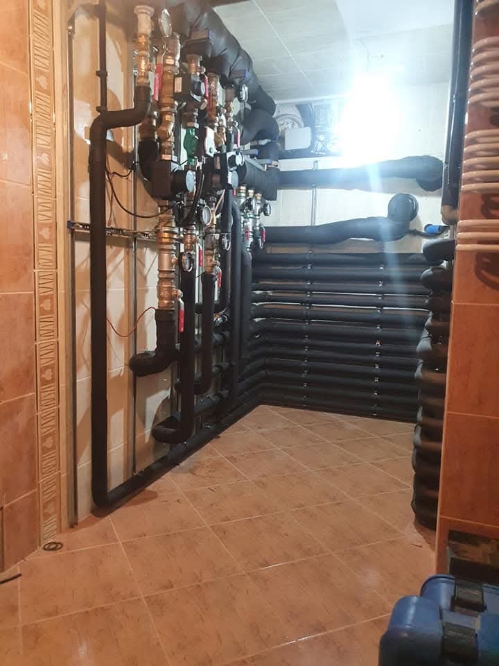 Rozbudowana instalacja centralnego ogrzewania z licznymi zaworami i rurami w izolacji termicznej, zamontowana w pomieszczeniu z wykończeniem ceramicznym.