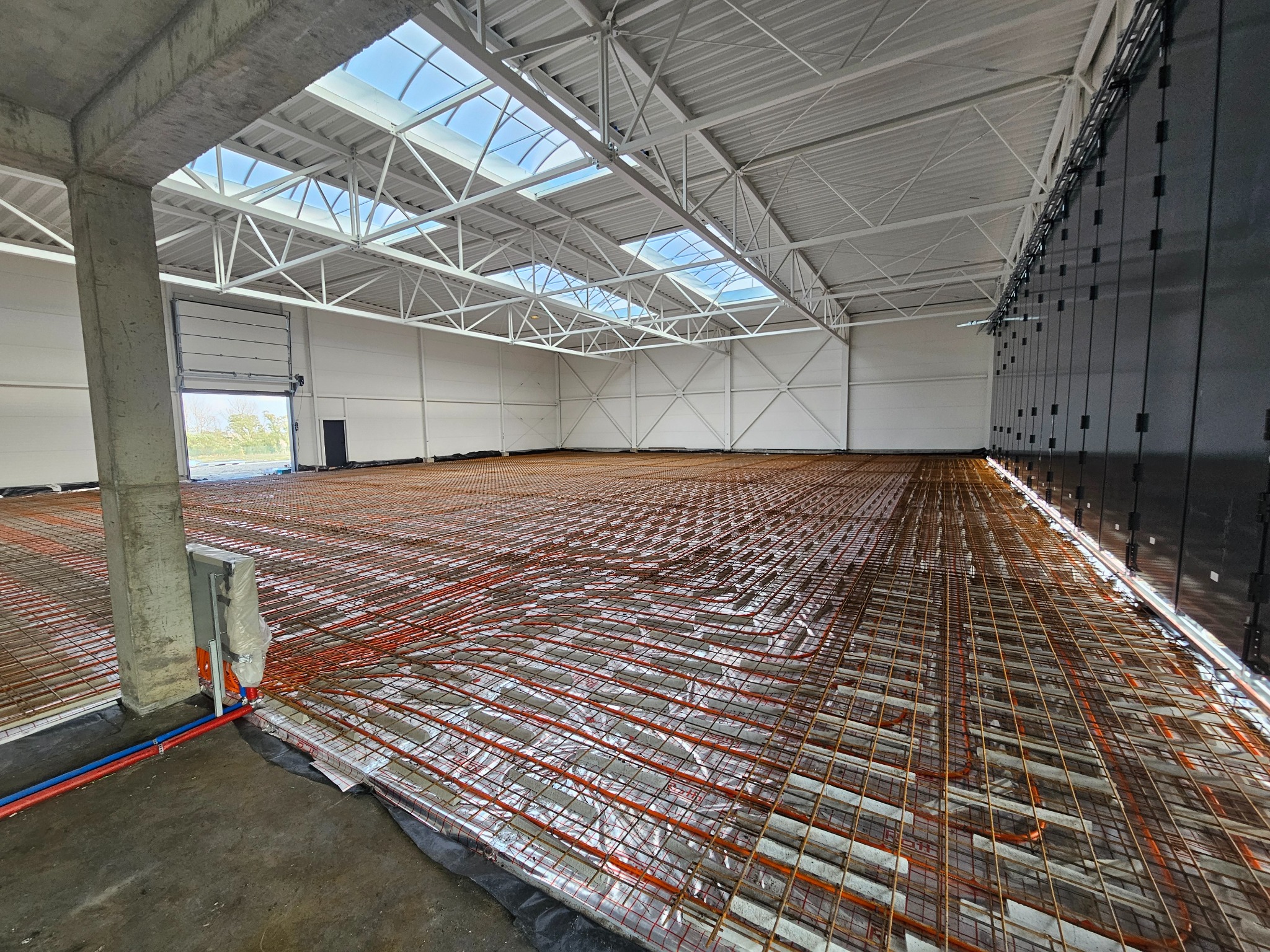 Rozległa hala przemysłowa z widoczną instalacją ogrzewania podłogowego w trakcie montażu, rury grzewcze ułożone na srebrnej folii izolacyjnej, betonowy filar w lewym dolnym rogu, błękitne...