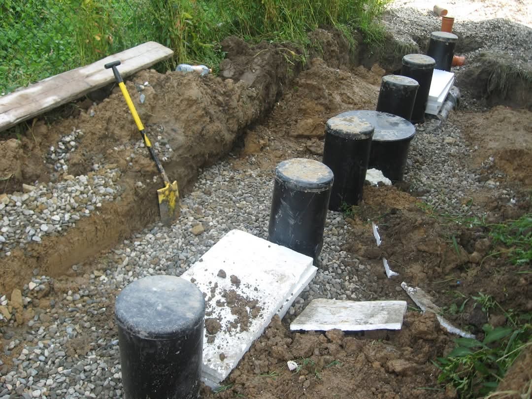 Wykop z ułożonym drenażem, czarne studzienki rozsączające i żółta łopata oparta o brzeg wykopu.