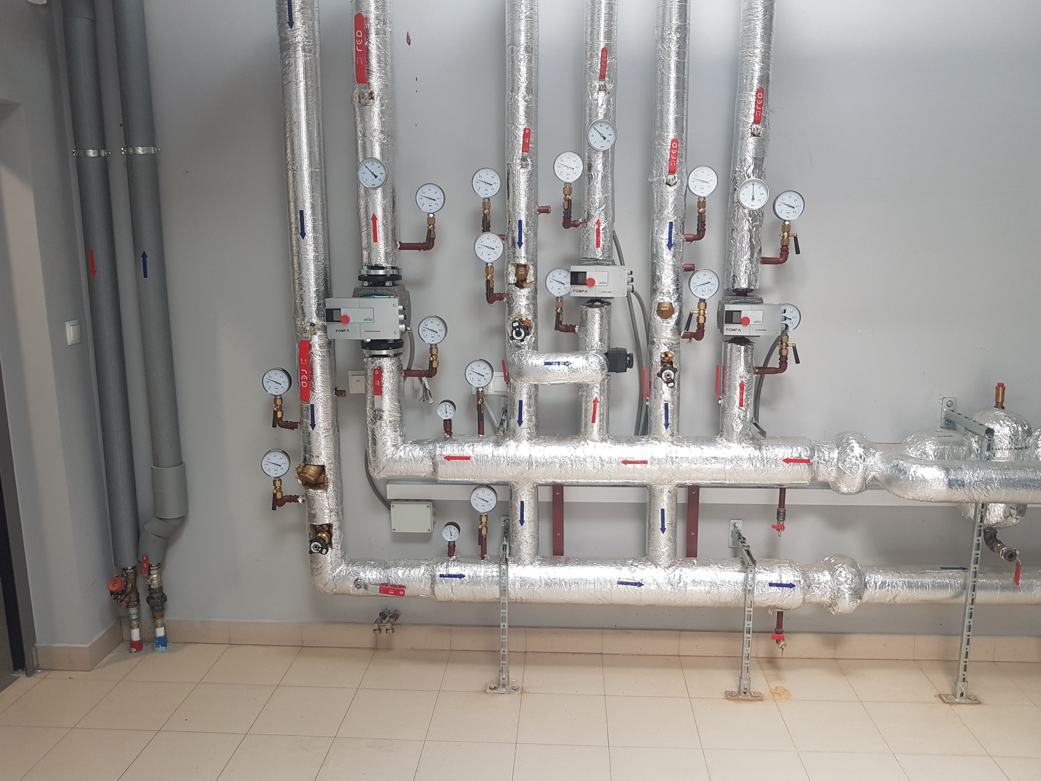 Izolowane rury instalacji grzewczej z manometrami, zaworami i oznaczeniami przepływu, zamontowane na ścianie w pomieszczeniu technicznym.