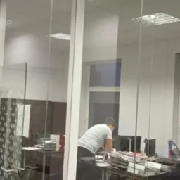 Przeszklona ścianka działowa z białymi ramami w biurze, widoczna część biurka i pracownicy w tle.