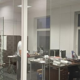 KAPA Usługi ogólnobudowlane Monika Stępińska - Szklana ścianka działowa w biurze z widocznym odkurzaczem piorącym i osobą w tle przy biurku.