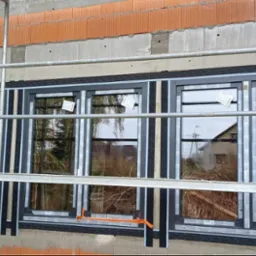 Szare ramy okienne w trakcie montażu w surowym stanie budynku, widoczne rusztowania, cegły i folia izolacyjna.