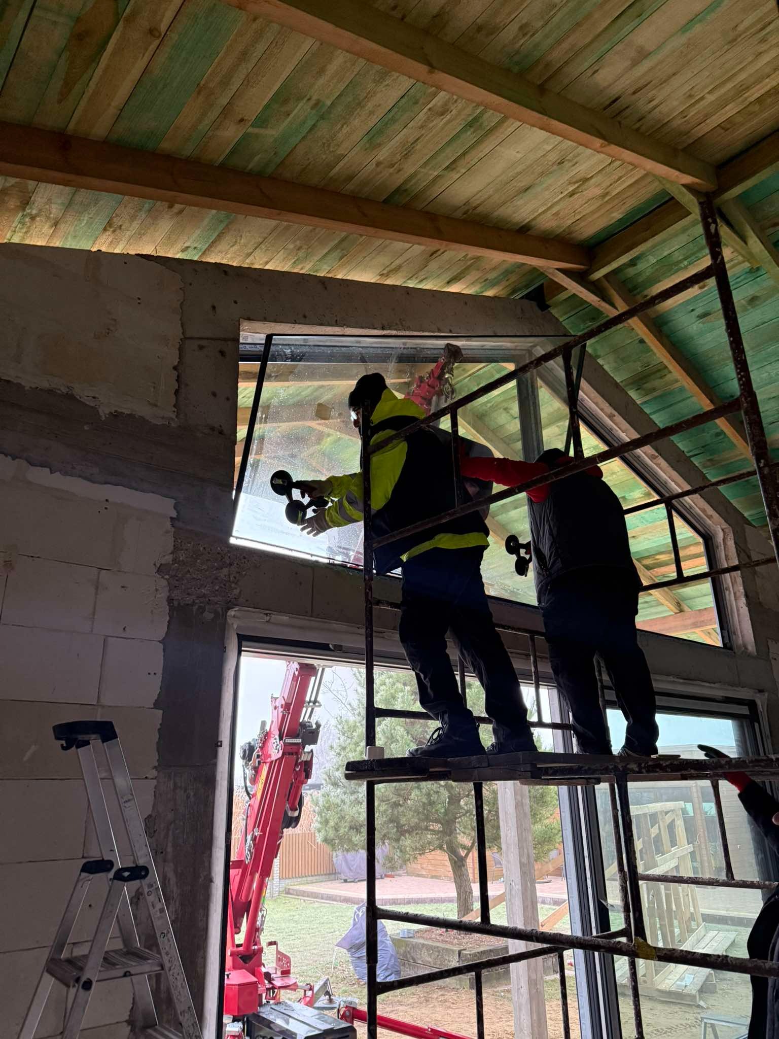 Dwóch monterów na rusztowaniu instaluje okno za pomocą przyssawek, widoczny podnośnik okienny na zewnątrz budynku, surowy stan wykończenia wnętrza.