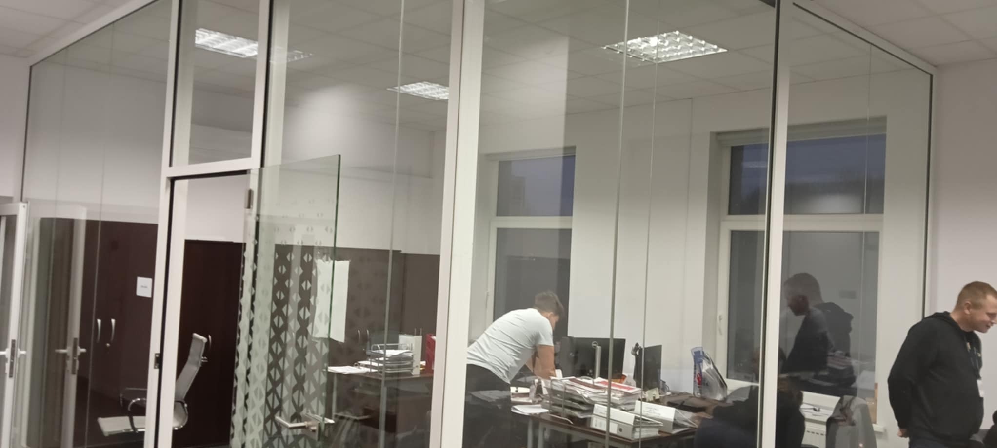Przeszklona ścianka działowa z białymi ramami w biurze, widoczna część biurka i pracownicy w tle.