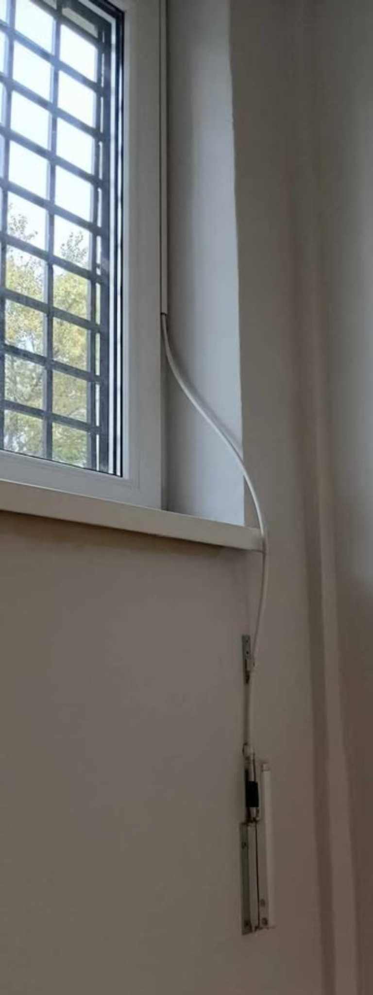Białe okno PCV z kratą zabezpieczającą, widoczny element okucia z linką sterującą zamontowany na ścianie wewnątrz pomieszczenia.