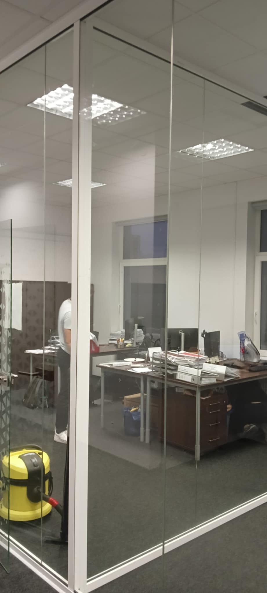 Szklana ścianka działowa w biurze z widocznym odkurzaczem piorącym i osobą w tle przy biurku.