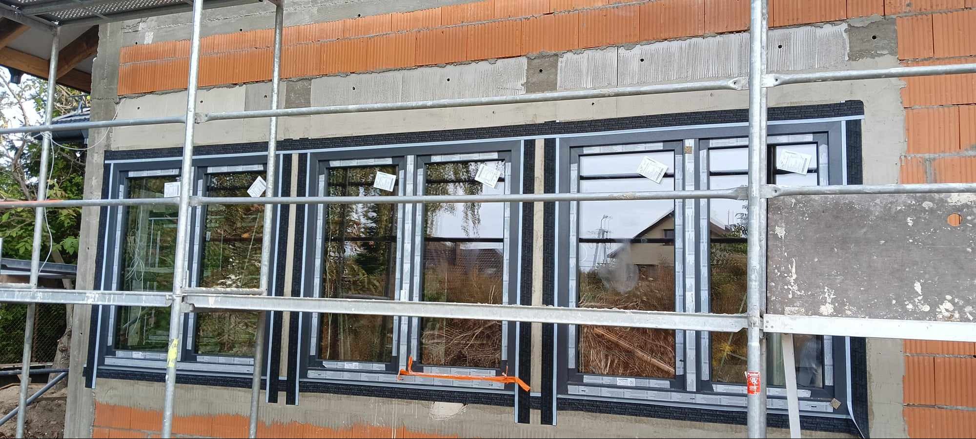 Szare ramy okienne w trakcie montażu w surowym stanie budynku, widoczne rusztowania, cegły i folia izolacyjna.