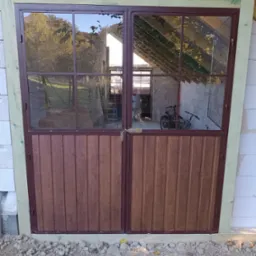 Dwuskrzydłowe, brązowe drzwi z metalową ramą i szybami, zamontowane w surowym stanie, widoczne elementy konstrukcyjne budynku, w tle rowery.