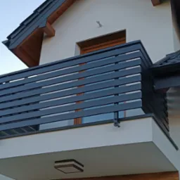 Nowoczesna, pozioma balustrada balkonowa w kolorze grafitowym, zamontowana na jasnym budynku z ciemnym dachem, widoczna z dołu pod kątem.