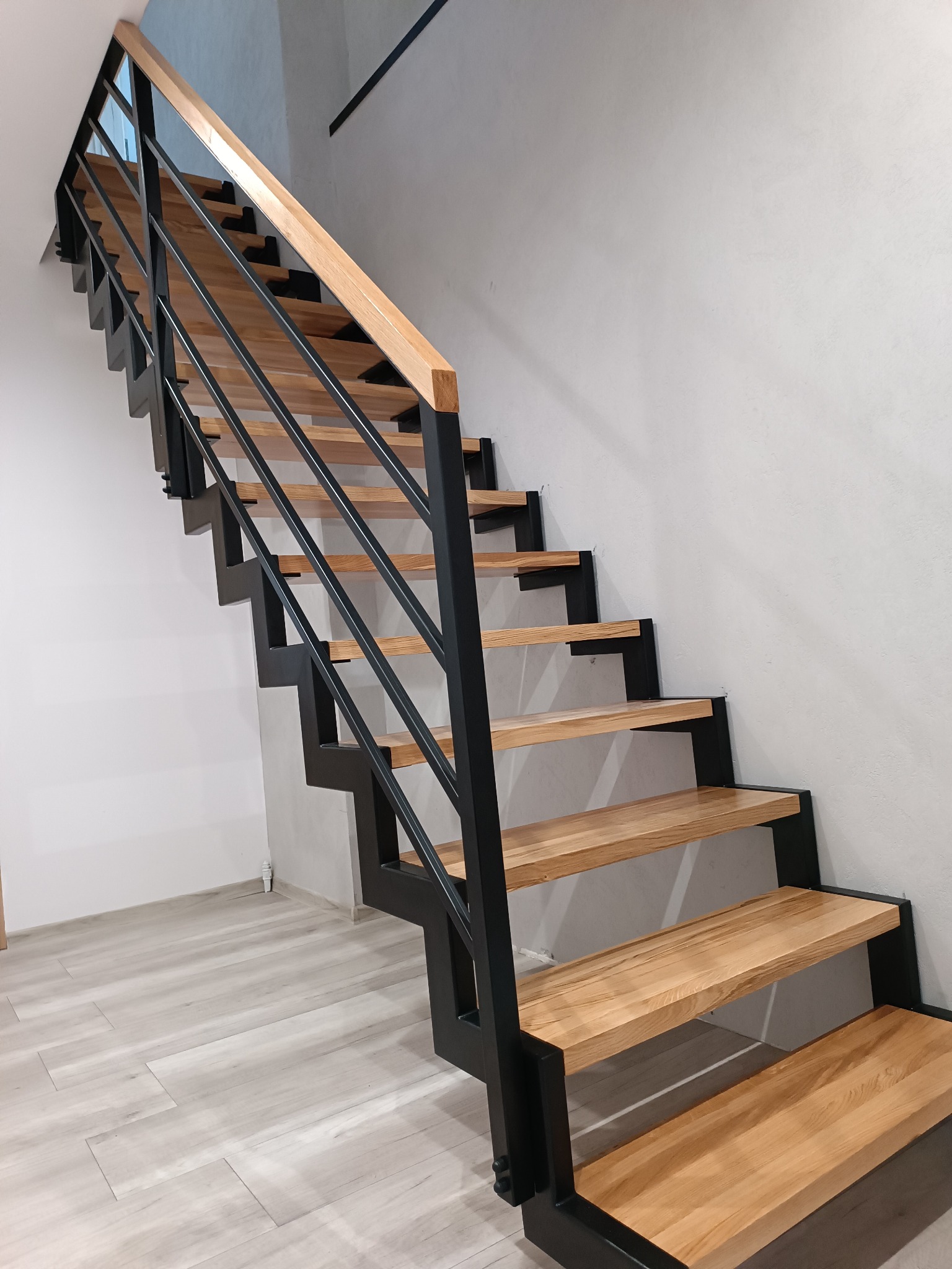 Nowoczesne schody z drewnianymi stopniami i czarną metalową konstrukcją oraz minimalistyczną balustradą z jasnego drewna, widok z dołu.