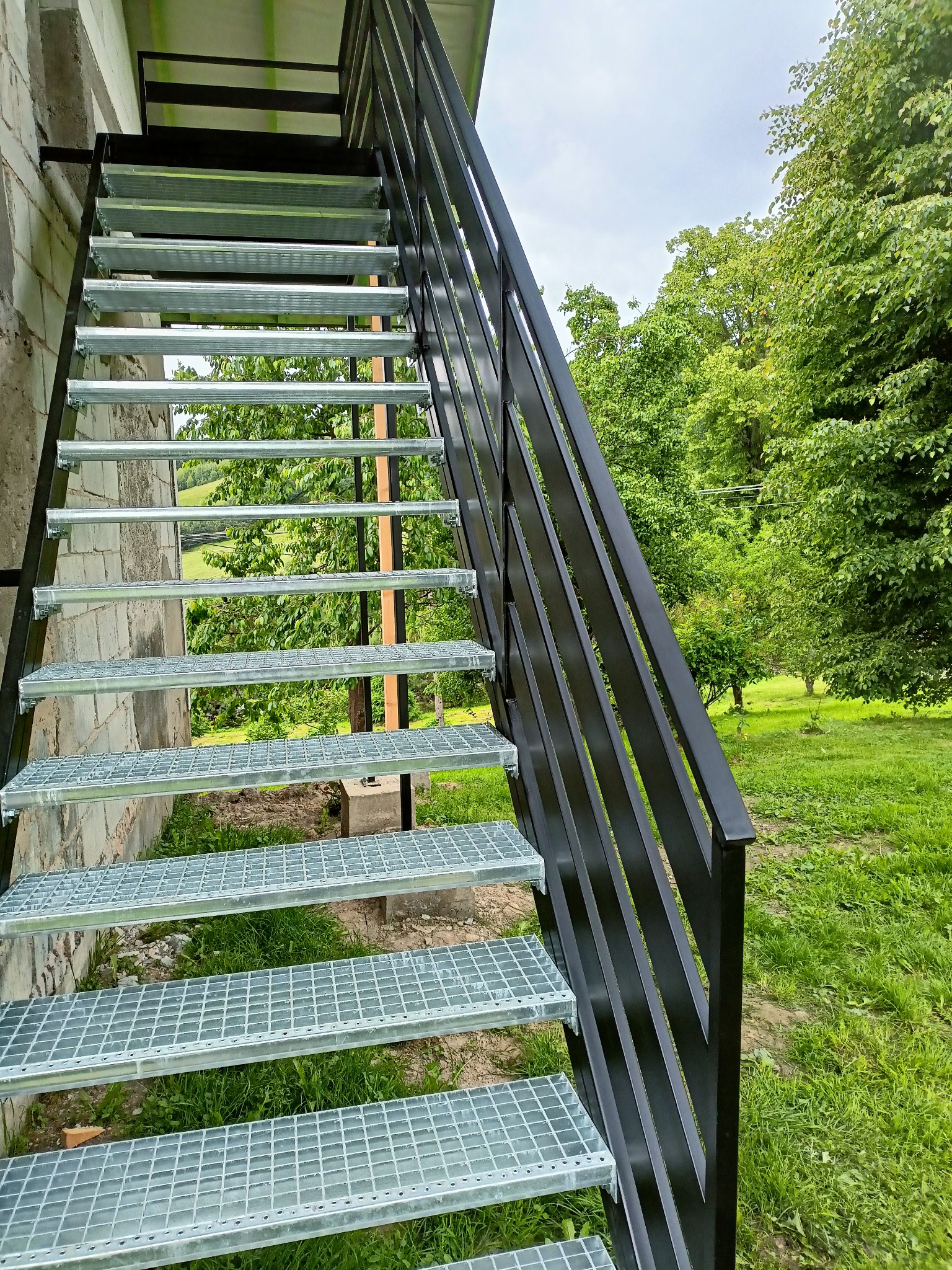 Zewnętrzne schody z metalową konstrukcją i kratowymi stopniami w Sanoku, prowadzące na wyższą kondygnację budynku, z widokiem na zielony ogród w tle.