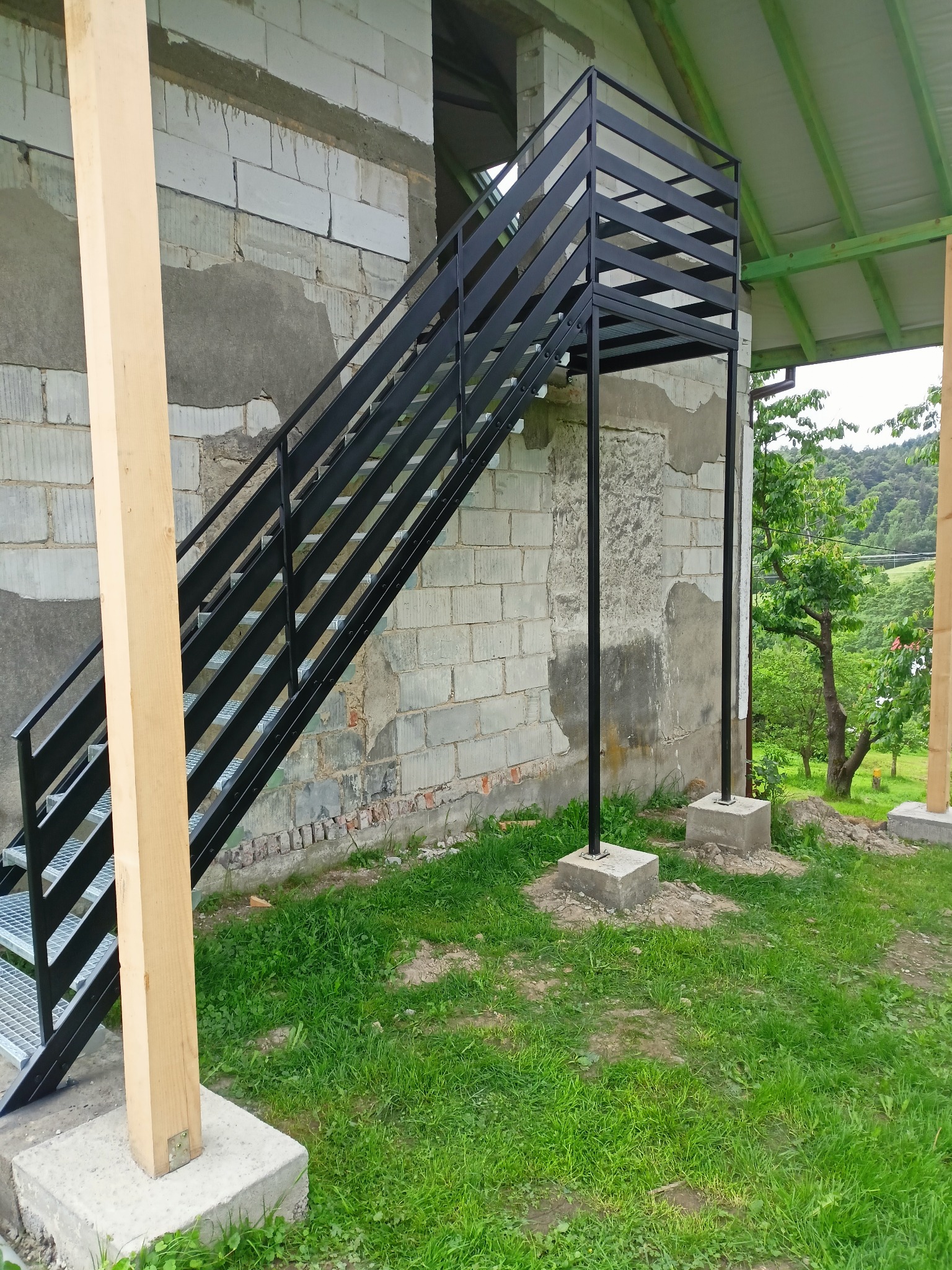 Zewnętrzne czarne schody z metalu z ażurowymi stopniami prowadzące do budynku w trakcie budowy, z widocznym drewnianym słupem i zielonym otoczeniem.
