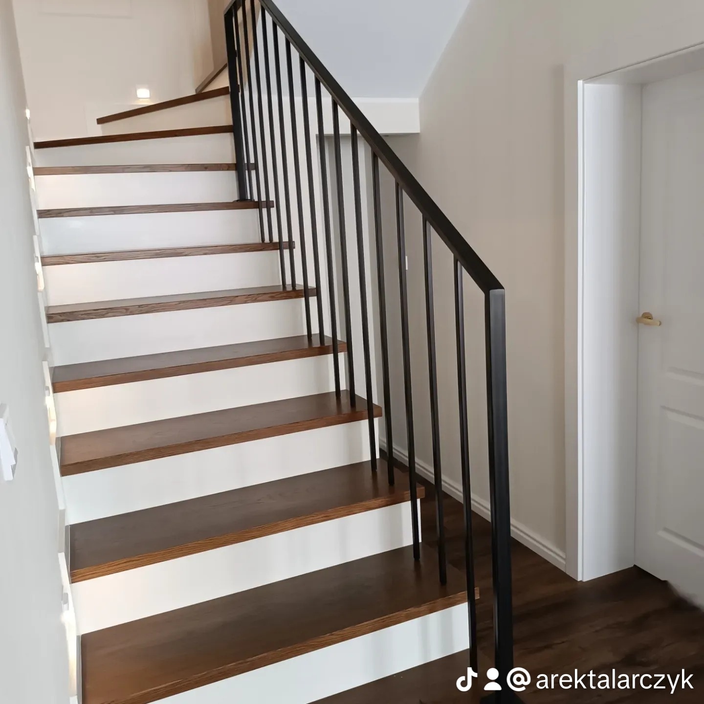 Nowoczesne schody drewniane z białymi podstopnicami i czarną metalową balustradą w jasnym wnętrzu, widok z dołu, minimalistyczny design.