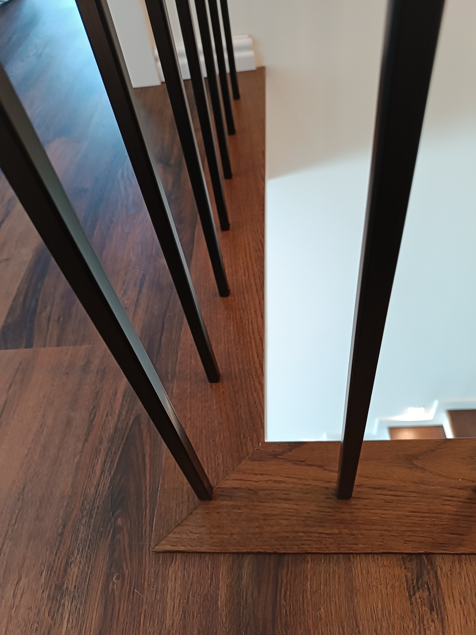 Fragment schodów z drewnianymi stopniami i czarnymi, pionowymi prętami balustrady, widoczne z bliskiej perspektywy, ukazujące detale wykończenia i teksturę drewna.