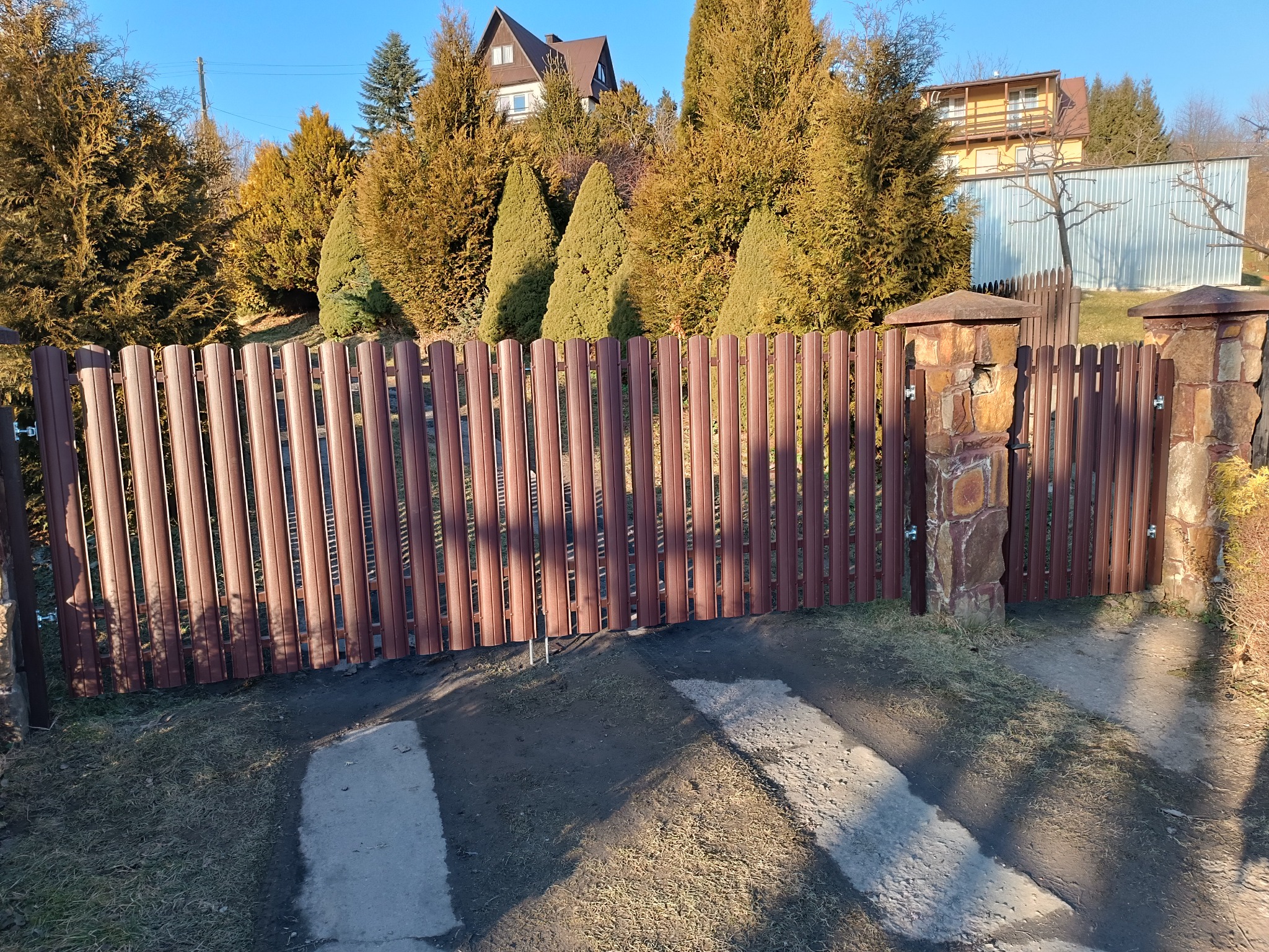 Brązowa brama z pionowych profili, osadzona na kamiennych słupkach, prowadząca do posesji z widocznymi domami i tujami w tle.