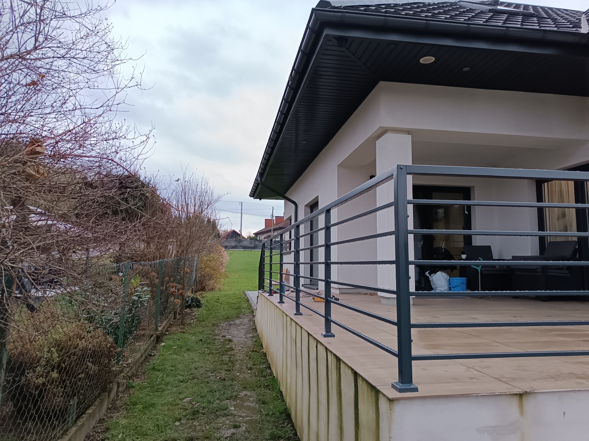 Nowoczesna, pozioma balustrada z ciemnego metalu otaczająca taras domu jednorodzinnego, widoczny zarośnięty trawnik i siatka ogrodzeniowa w tle, pochmurne niebo.