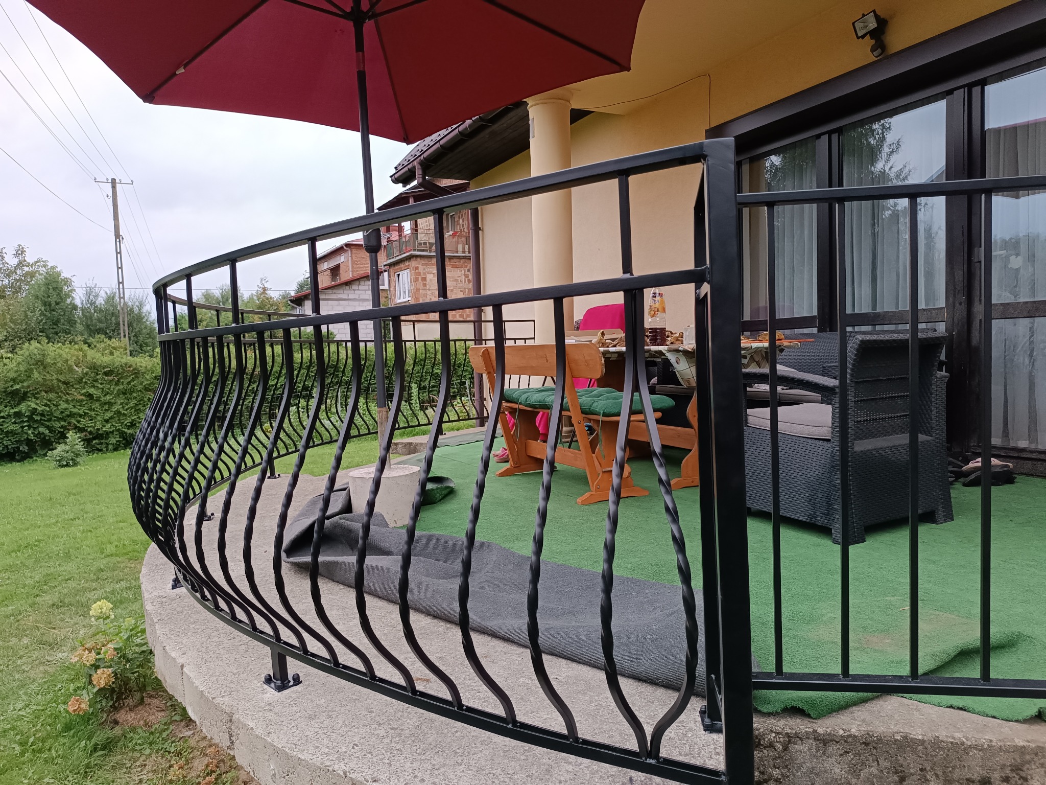 Czarna, metalowa balustrada balkonowa z ozdobnymi, skręconymi elementami, widok na zielony trawnik i zabudowę mieszkalną w tle, częściowo zasłonięta czerwonym parasolem.