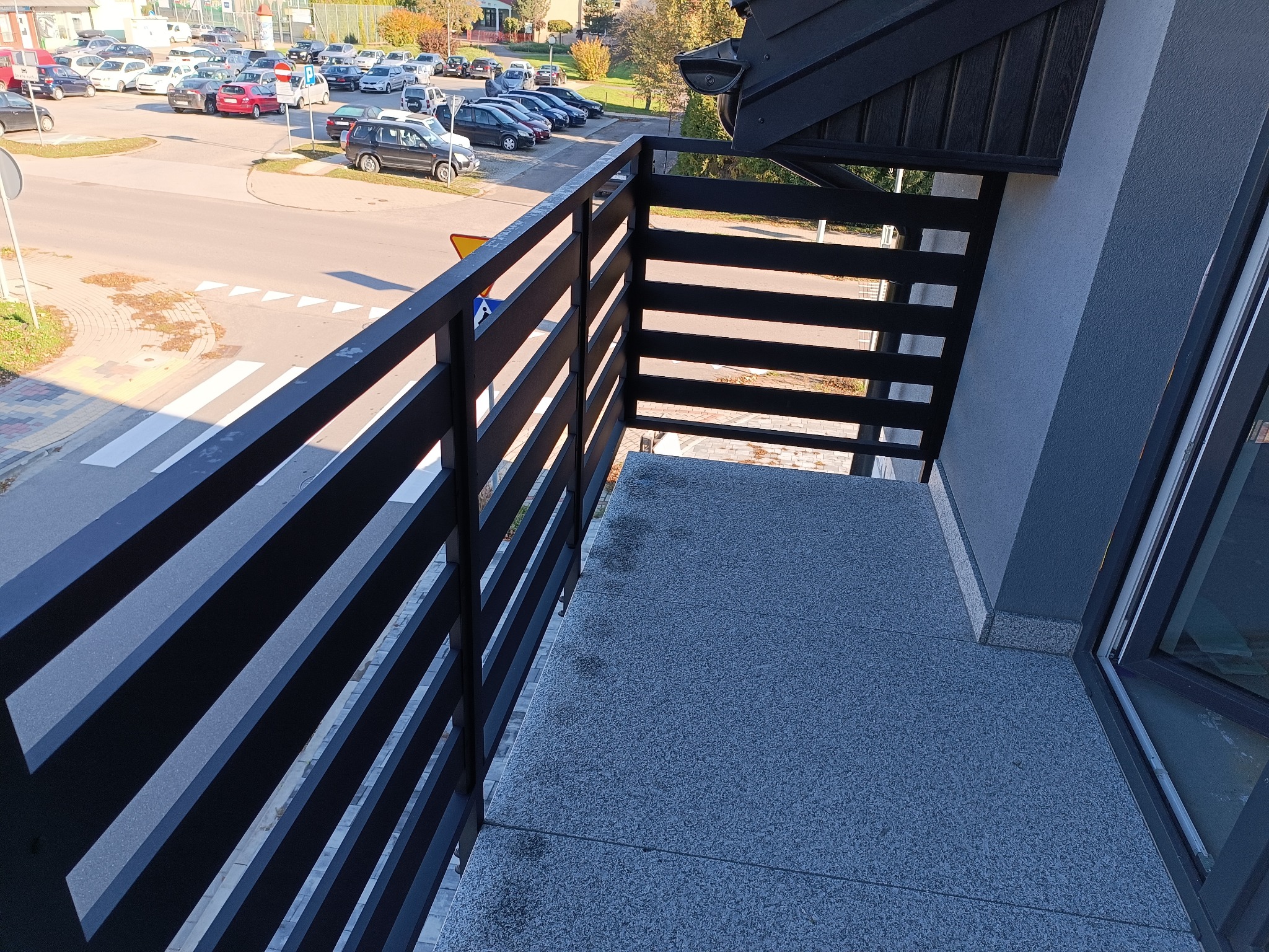 Nowoczesny balkon z czarną, poziomą balustradą, widok z góry na ulicę i parking w słoneczny dzień.