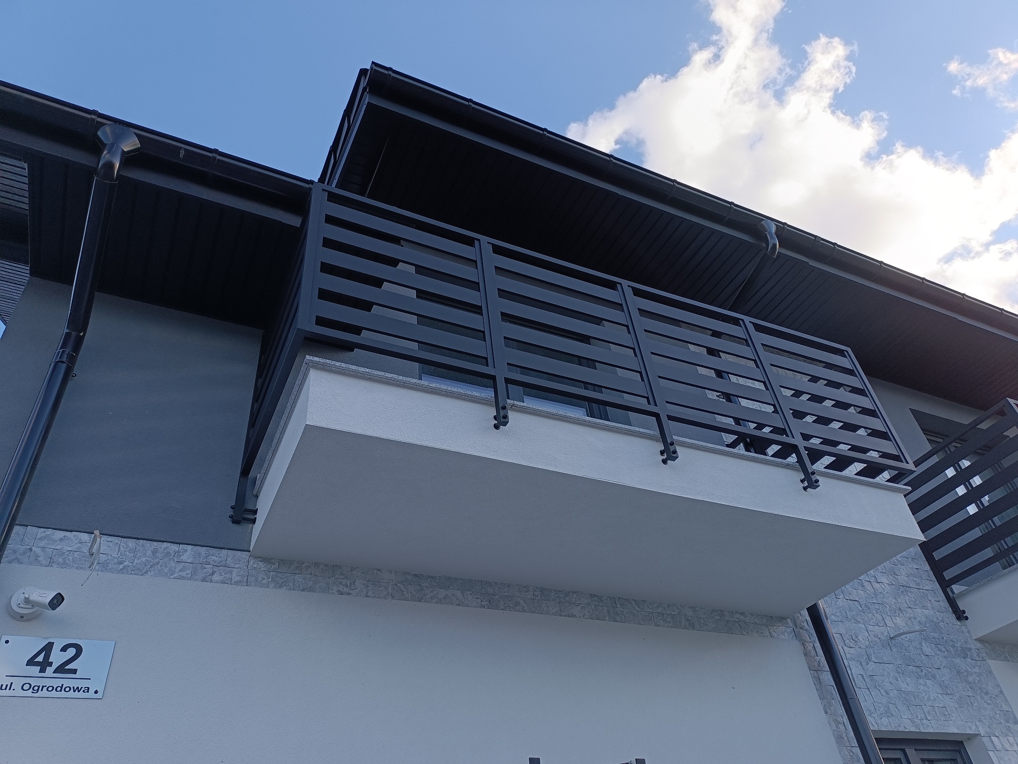 Nowoczesny balkon z czarną, poziomą balustradą, widok z dołu na elewację budynku z numerem 42 i błękitne niebo z chmurami.