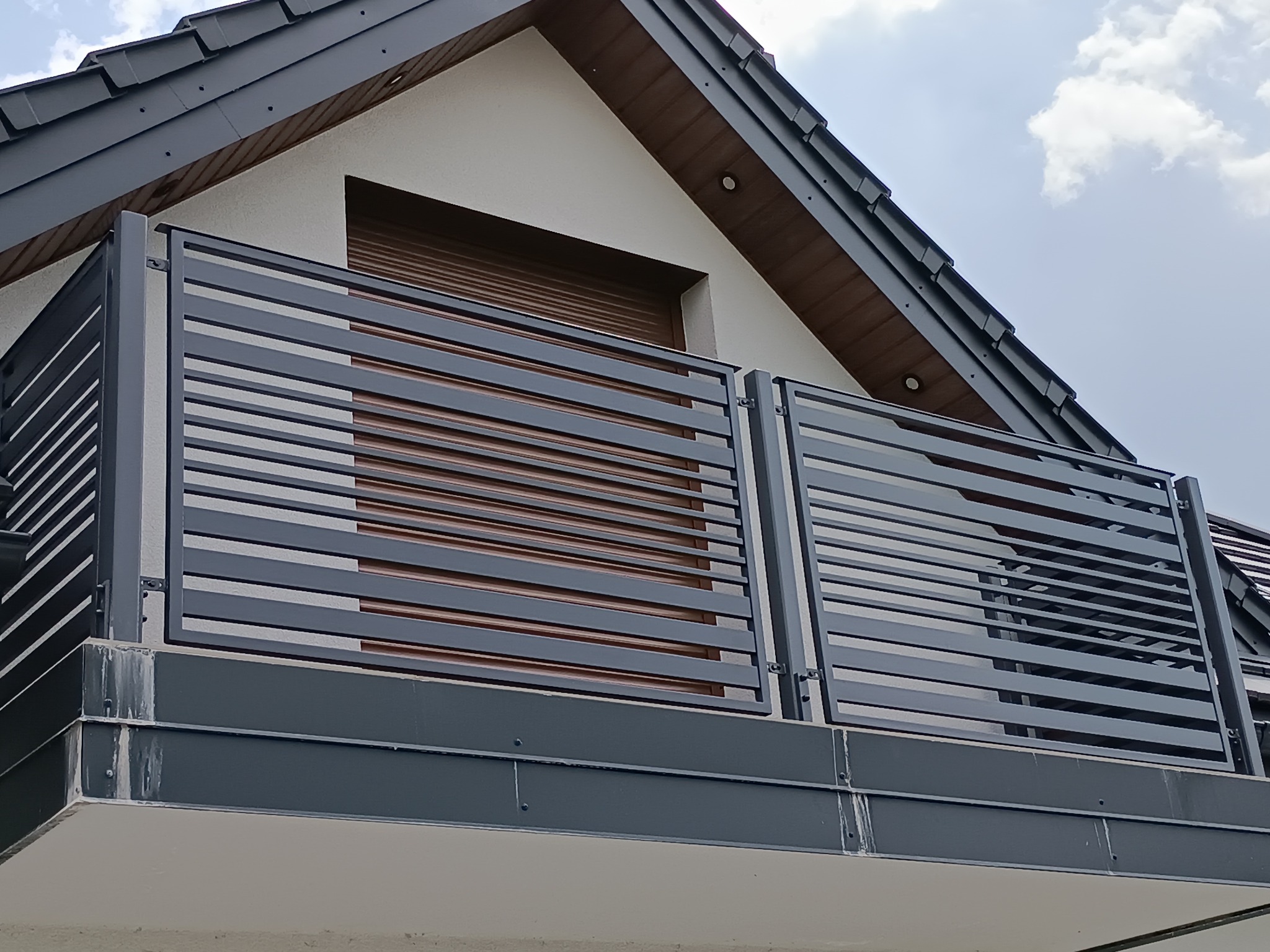 Nowoczesna, pozioma, metalowa balustrada balkonowa w kolorze grafitowym, z widocznym fragmentem elewacji domu i roletami okiennymi w tle.