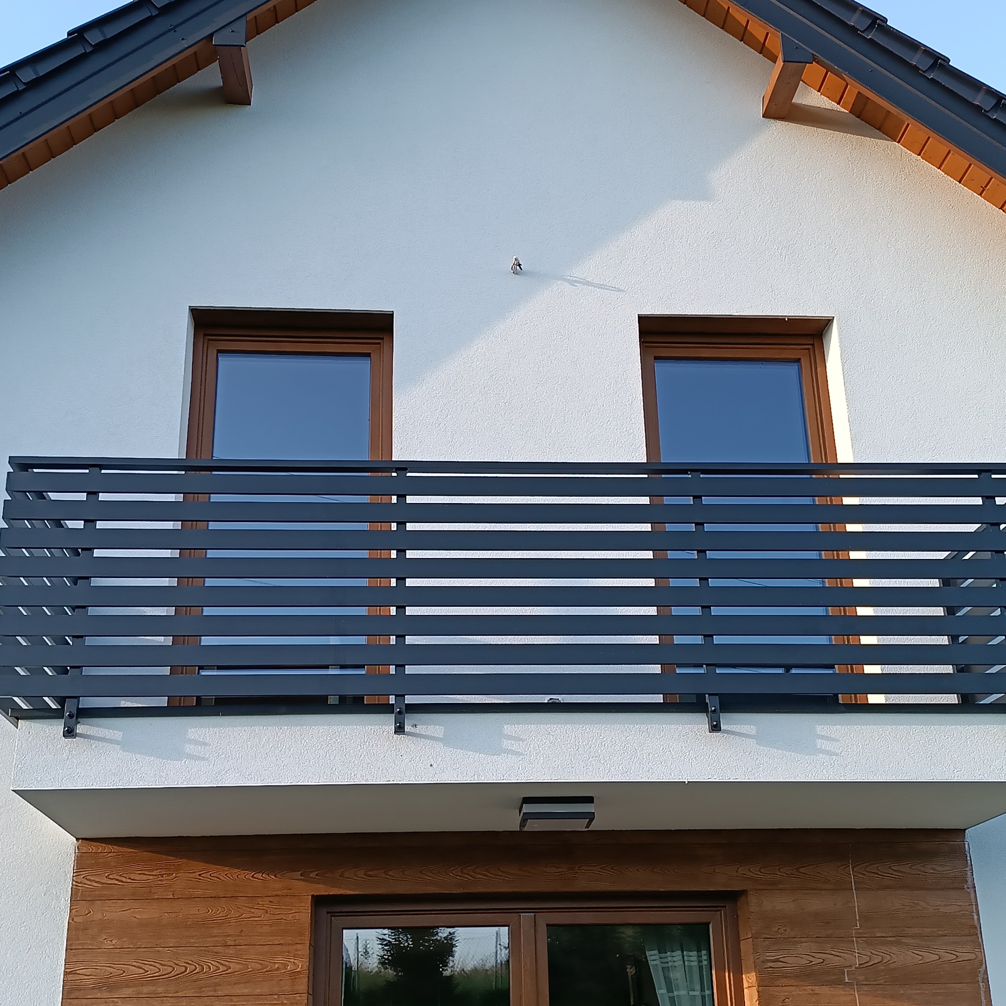 Nowoczesny balkon z poziomą, grafitową konstrukcją, widok z dołu na białą elewację domu z dwoma oknami w brązowych ramach, drewniana okładzina pod balkonem.