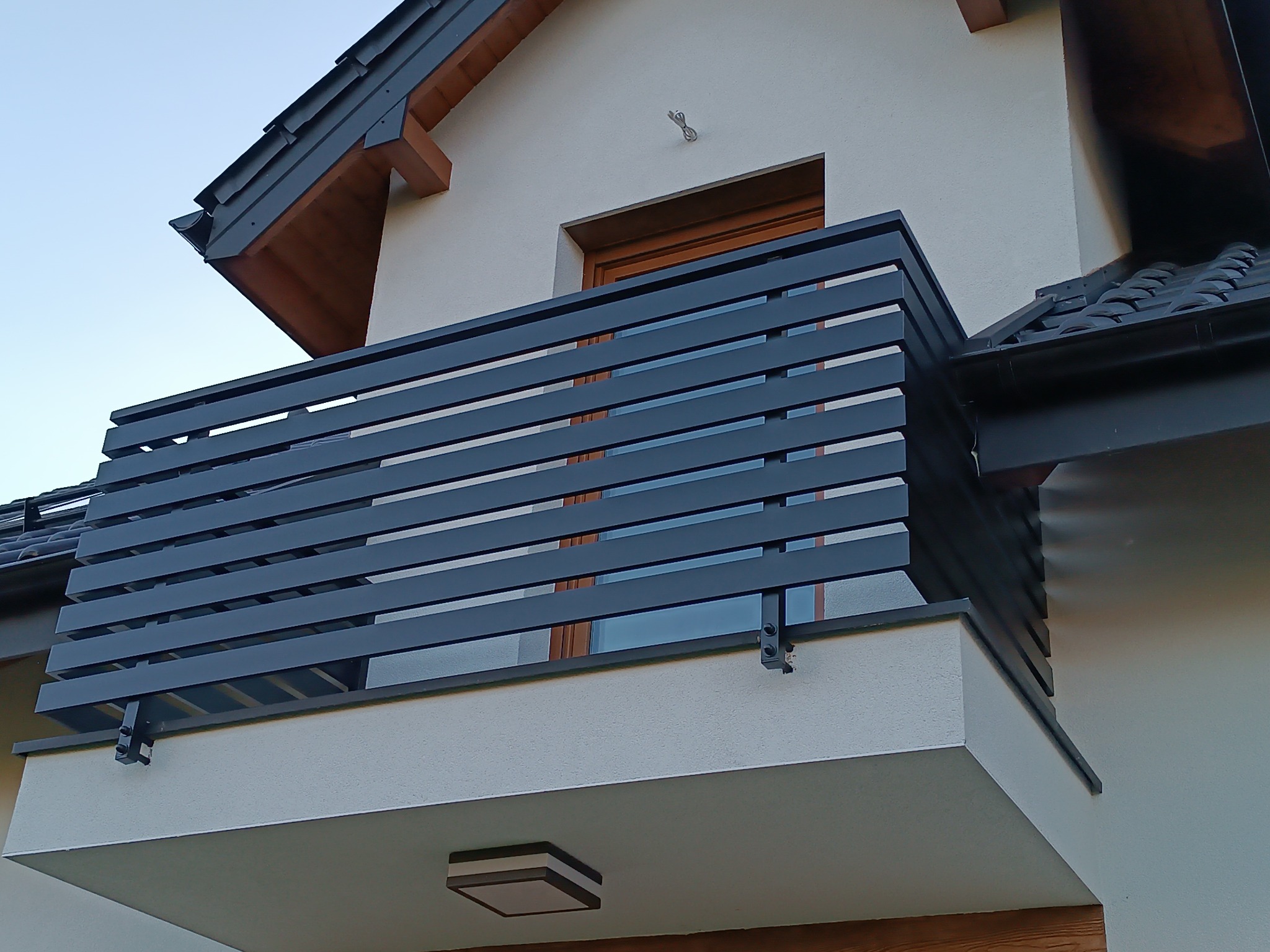 Nowoczesna, pozioma balustrada balkonowa w kolorze grafitowym, zamontowana na jasnym budynku z ciemnym dachem, widoczna z dołu pod kątem.