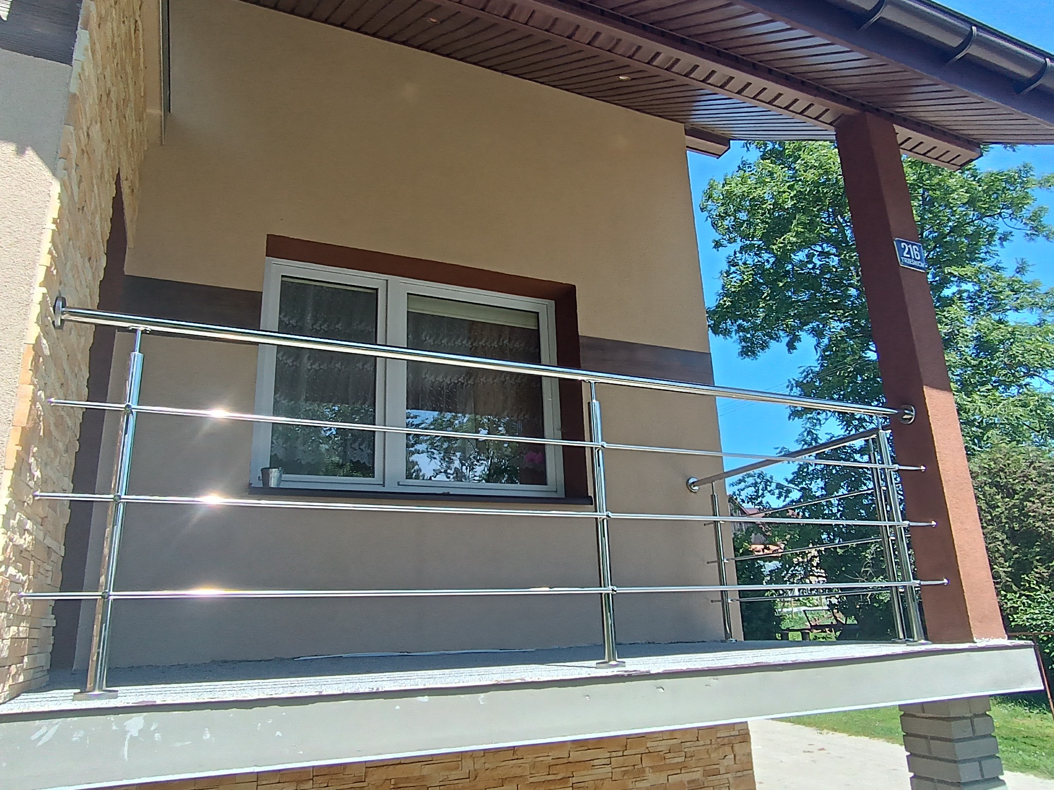 Błyszcząca, metalowa balustrada balkonowa z rurek, zamontowana przy oknie domu o beżowej elewacji, z elementami dekoracyjnymi z kamienia. Widok z dołu, na tle zieleni.