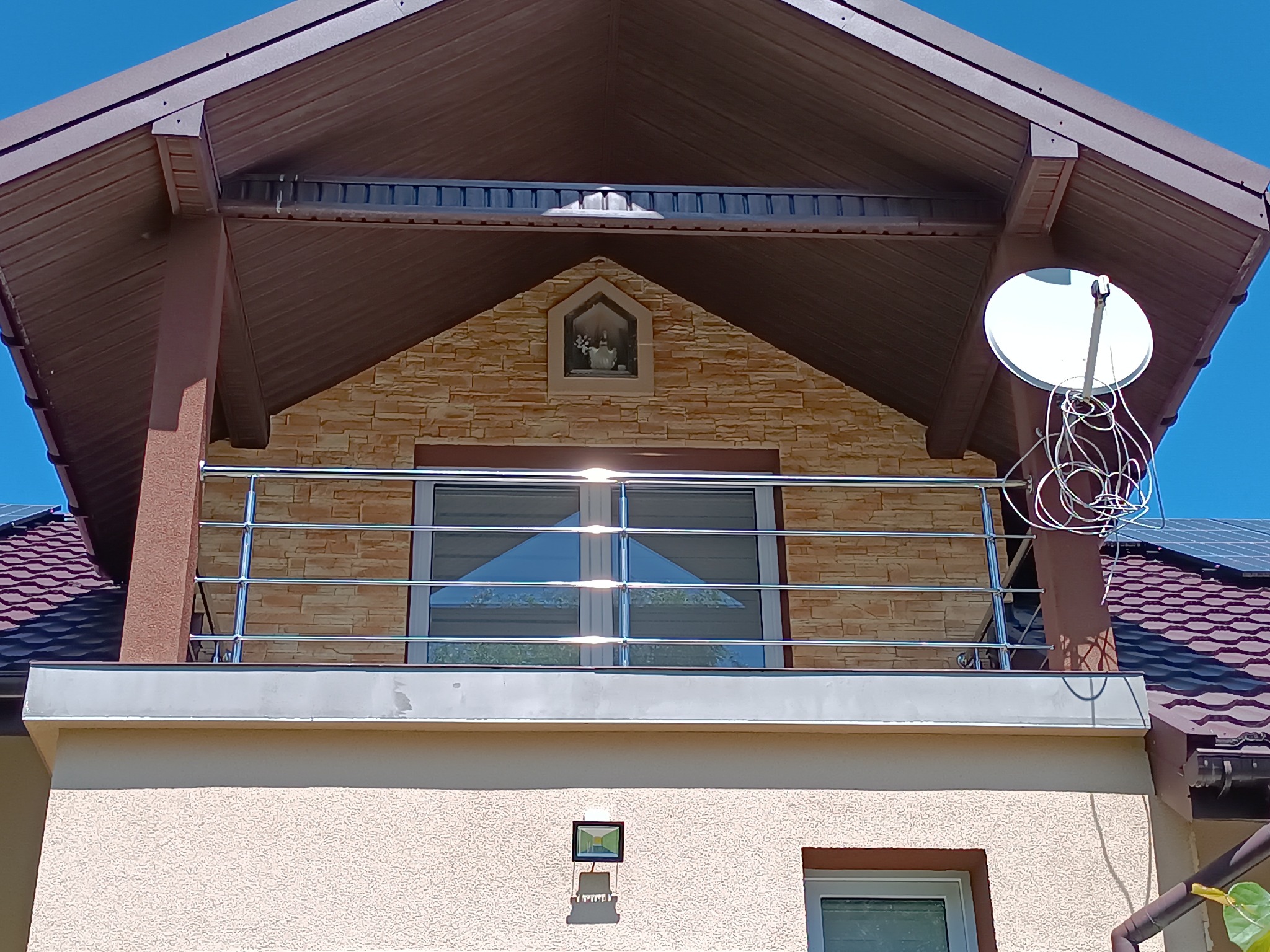 Balkon z metalową balustradą, widok z dołu, jasne światło słoneczne, brązowy dach, elewacja z dekoracyjną cegłą i małą kapliczką. Antena satelitarna na dachu.