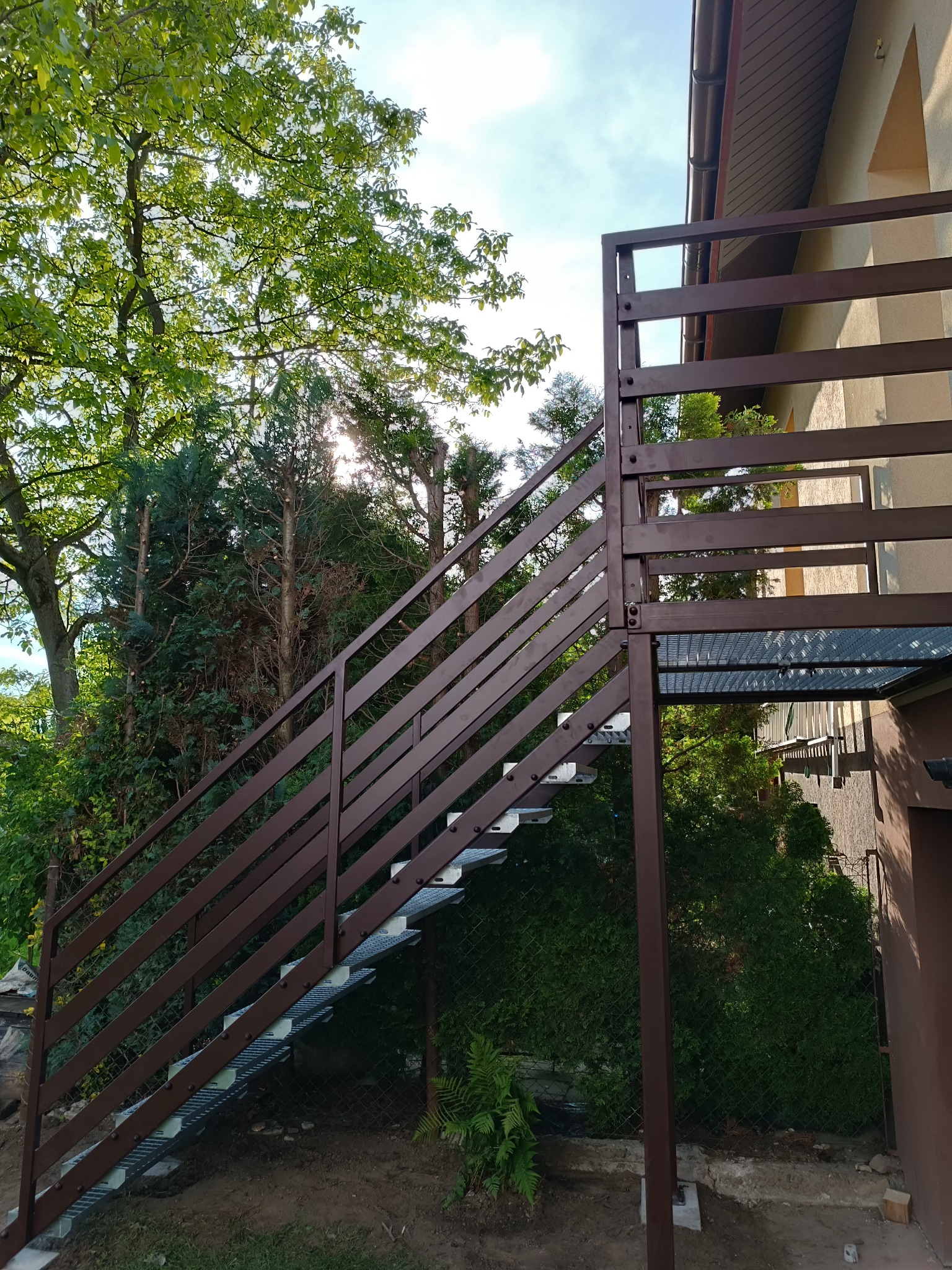 Zewnętrzne schody metalowe z brązową balustradą, prowadzące do balkonu domu. Widok z dołu, zieleń w tle. Schody z ażurowymi stopniami.