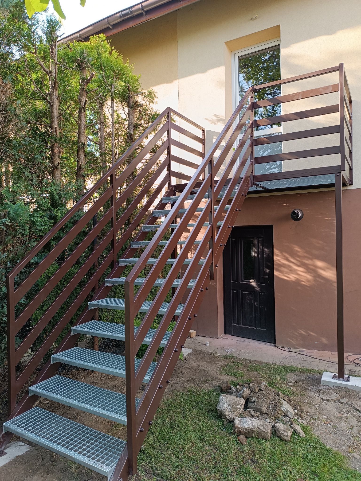 Zewnętrzne schody metalowe w kolorze brązowym prowadzące na balkon. Schody z kraty pomostowej, balustrada pozioma. Budynek w tle, zieleń, fragment kostki brukowej.