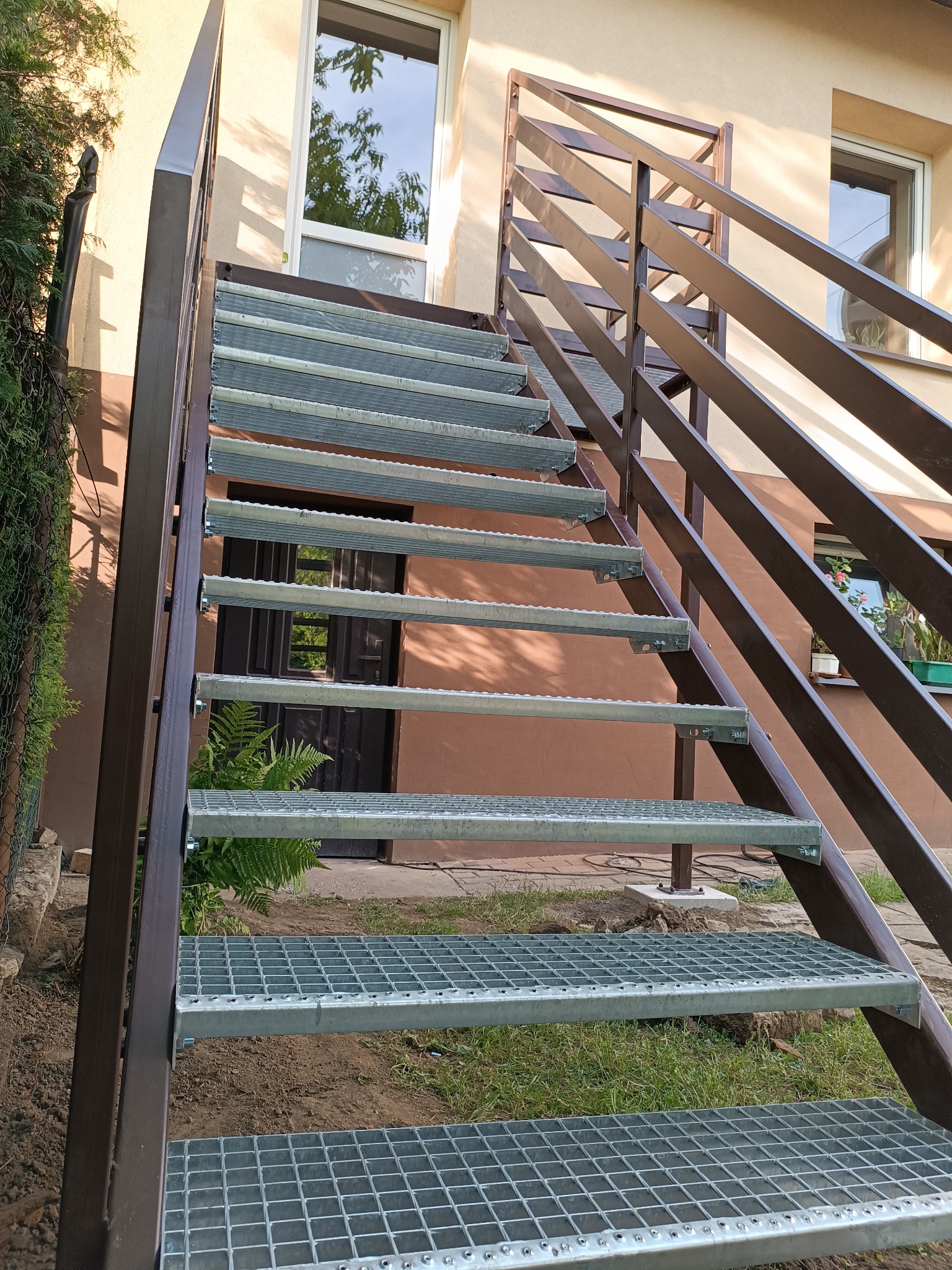 Zewnętrzne schody z metalową kratą i brązową balustradą, prowadzące do drzwi budynku. Widoczne rośliny w otoczeniu schodów.