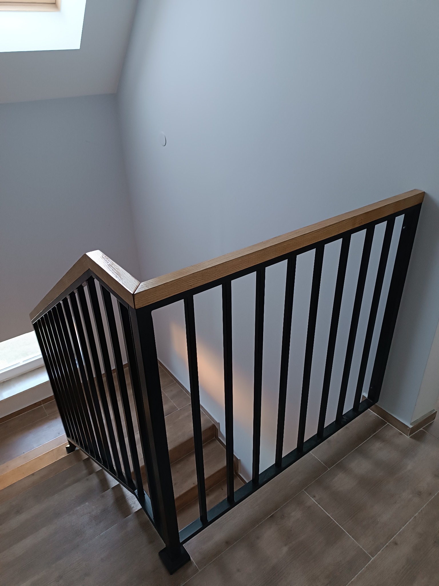 Nowoczesna balustrada schodowa z czarnymi, pionowymi prętami i drewnianym poręczą, widziana z góry, z oknem dachowym w tle. Minimalistyczny design.