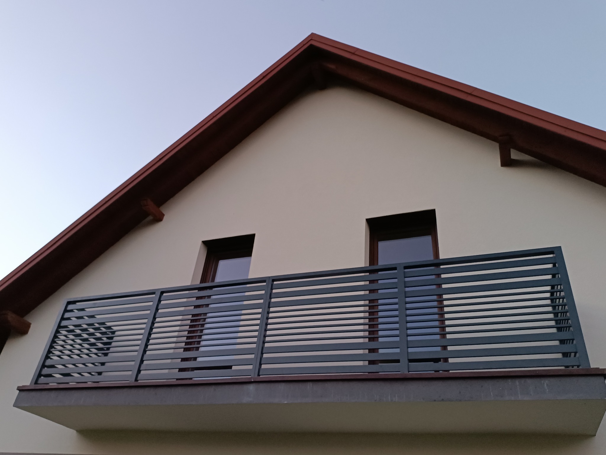 Nowoczesna balustrada balkonowa z poziomych profili, szara, na tle elewacji domu z brązowym dachem. Ujęcie z dołu, podkreślające geometrię i minimalistyczny design.