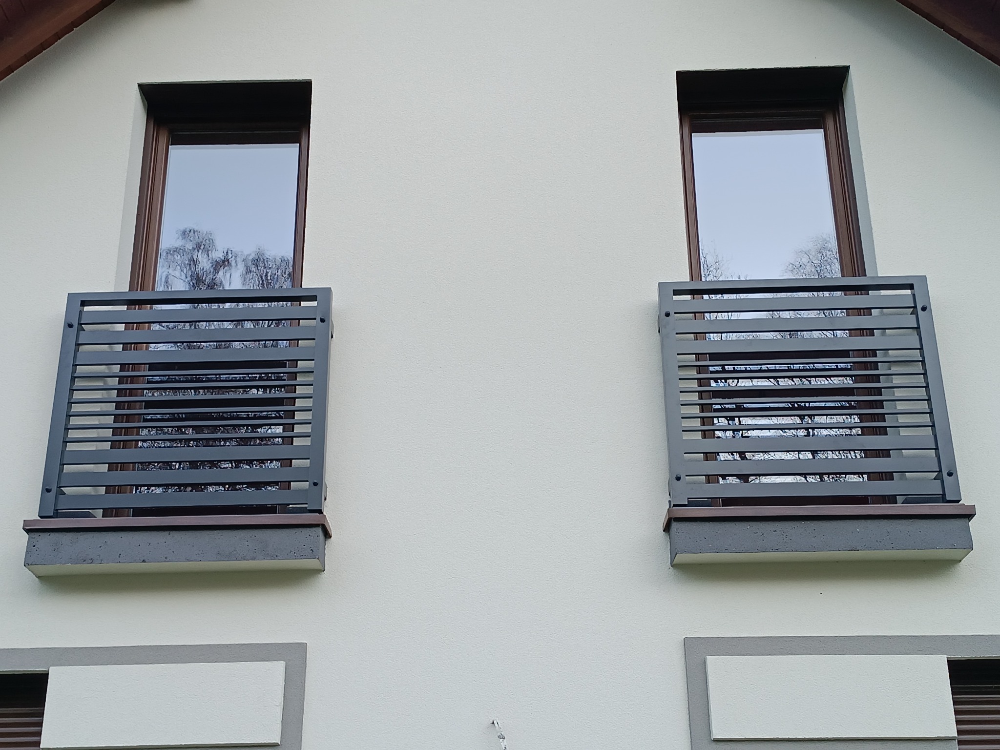 Dwa okna z nowoczesnymi, poziomymi, metalowymi osłonami w kolorze grafitowym, zamontowanymi na betonowych parapetach. Widok na elewację budynku.