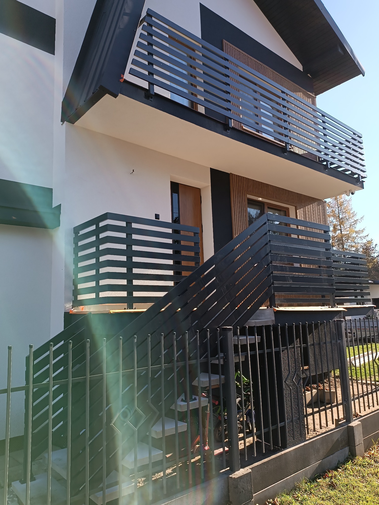 Nowoczesna, czarna balustrada balkonowa i schodowa z poziomymi szczeblami, dopasowana do elewacji domu z elementami drewnianymi. Widok z dołu, w tle błękitne niebo.