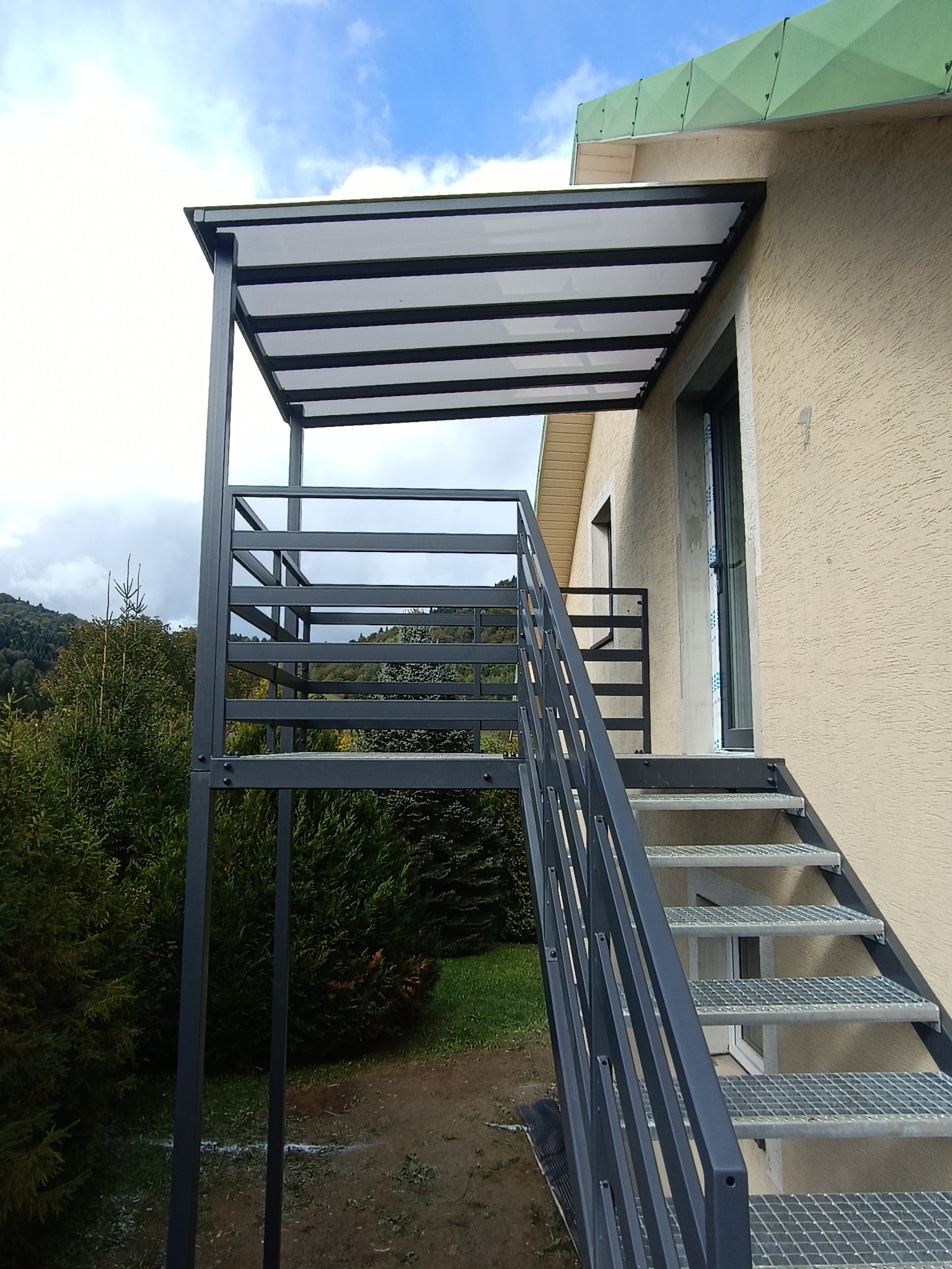 Metalowe schody zewnętrzne z zadaszeniem przy budynku o beżowej elewacji. Konstrukcja w kolorze grafitowym, ażurowe stopnie. Widok z dołu, błękitne niebo w tle.