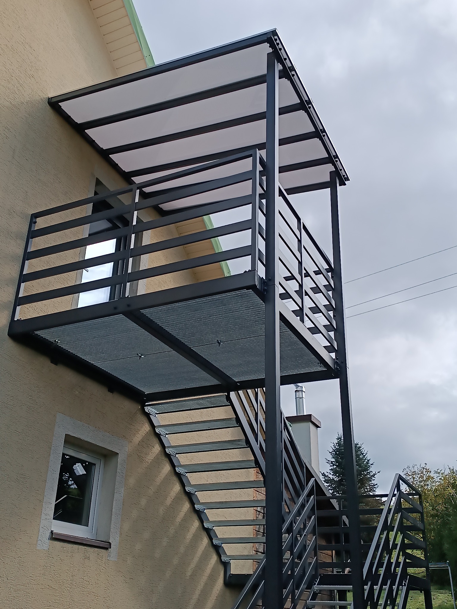 Zewnętrzne schody metalowe z balustradą i zadaszeniem, prowadzące na balkon. Konstrukcja w kolorze grafitowym, widok z dołu, na tle beżowej elewacji budynku.
