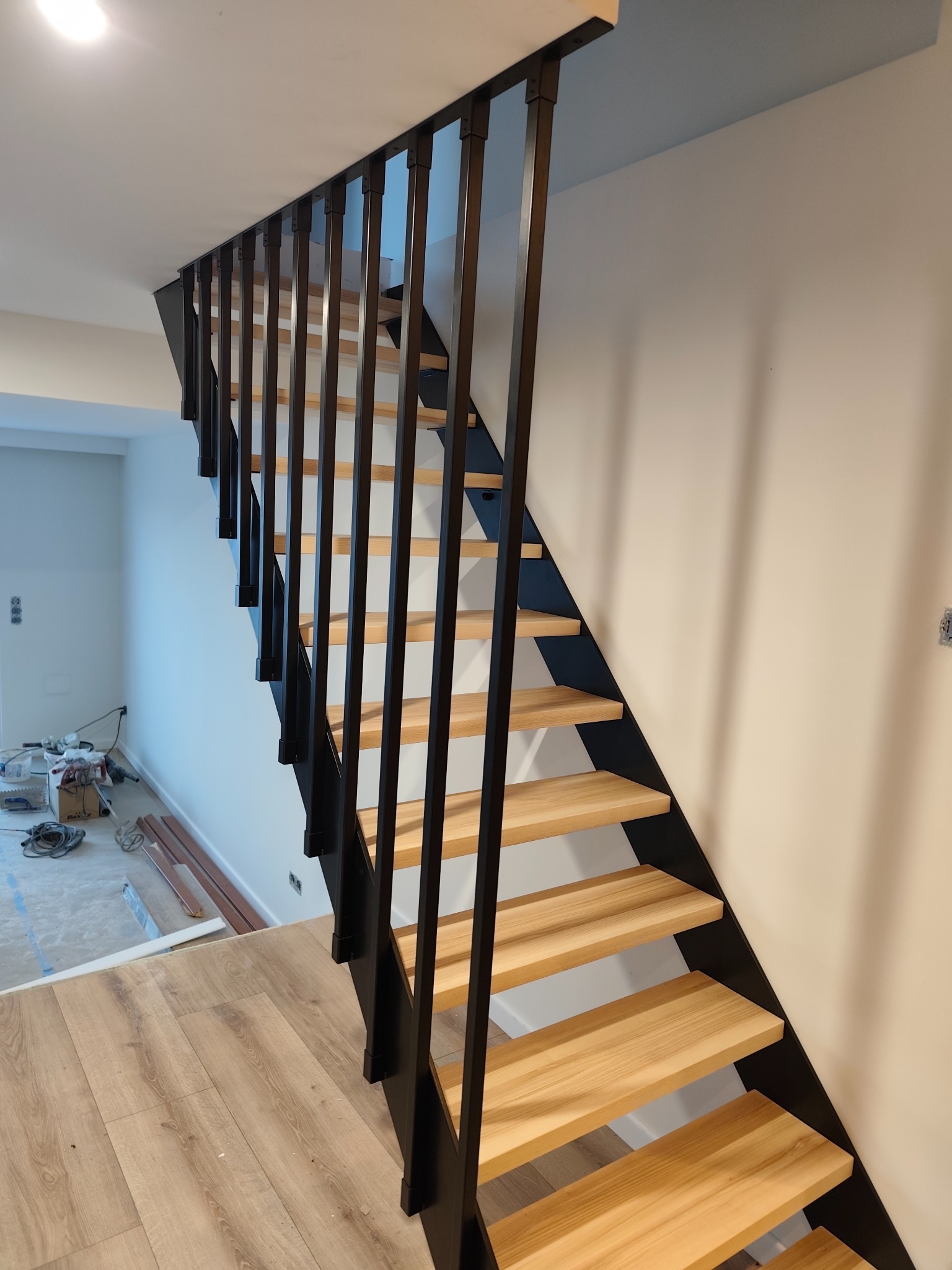 Nowoczesne schody z drewnianymi stopniami i czarną, metalową balustradą. Minimalistyczny design, jasne wnętrze w trakcie wykończenia. Widok z dołu na górę.