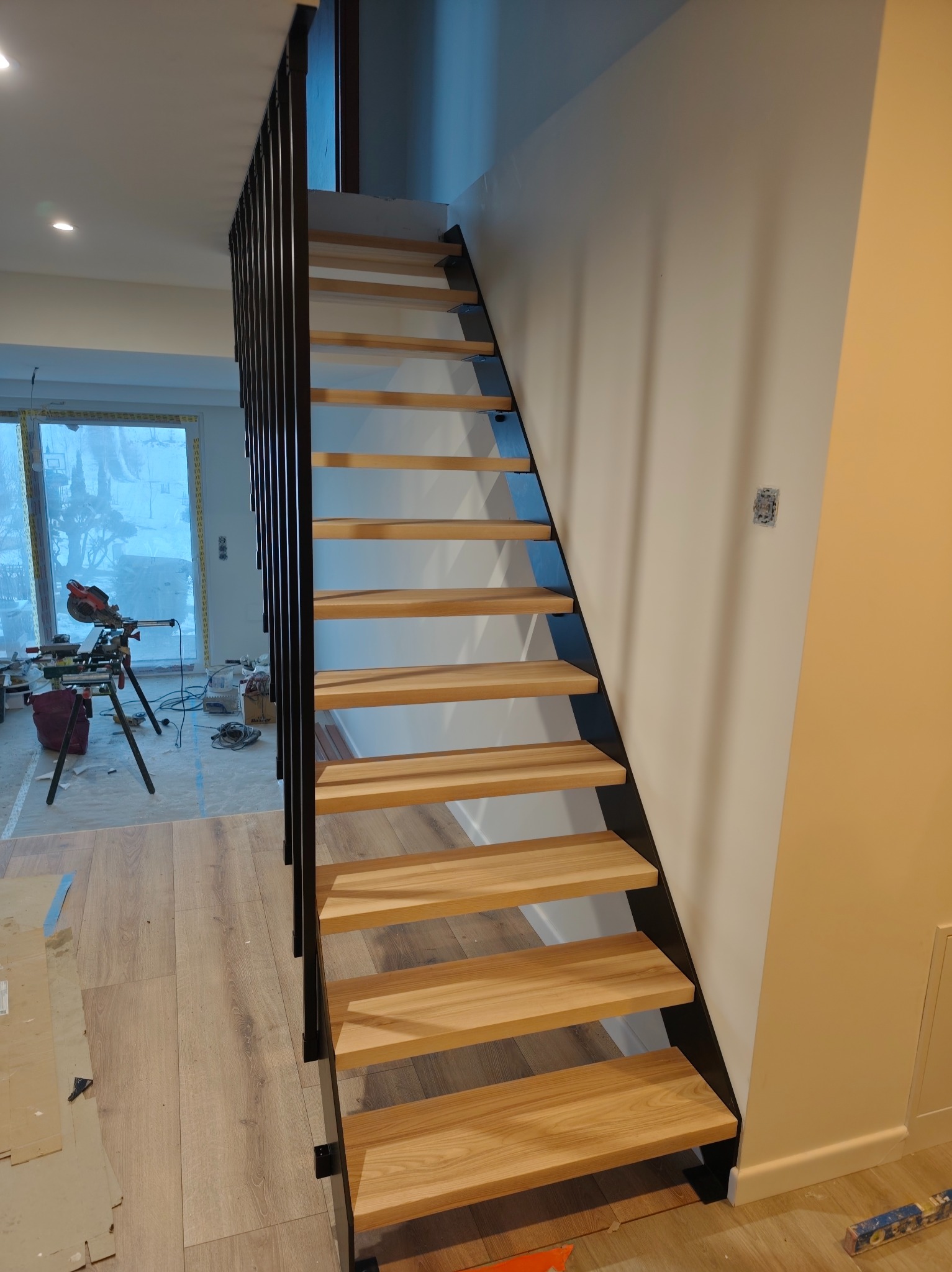 Nowoczesne schody z drewnianymi stopniami i czarną metalową balustradą. W tle widoczny sprzęt budowlany, co sugeruje etap montażu lub świeżo zakończoną instalację.