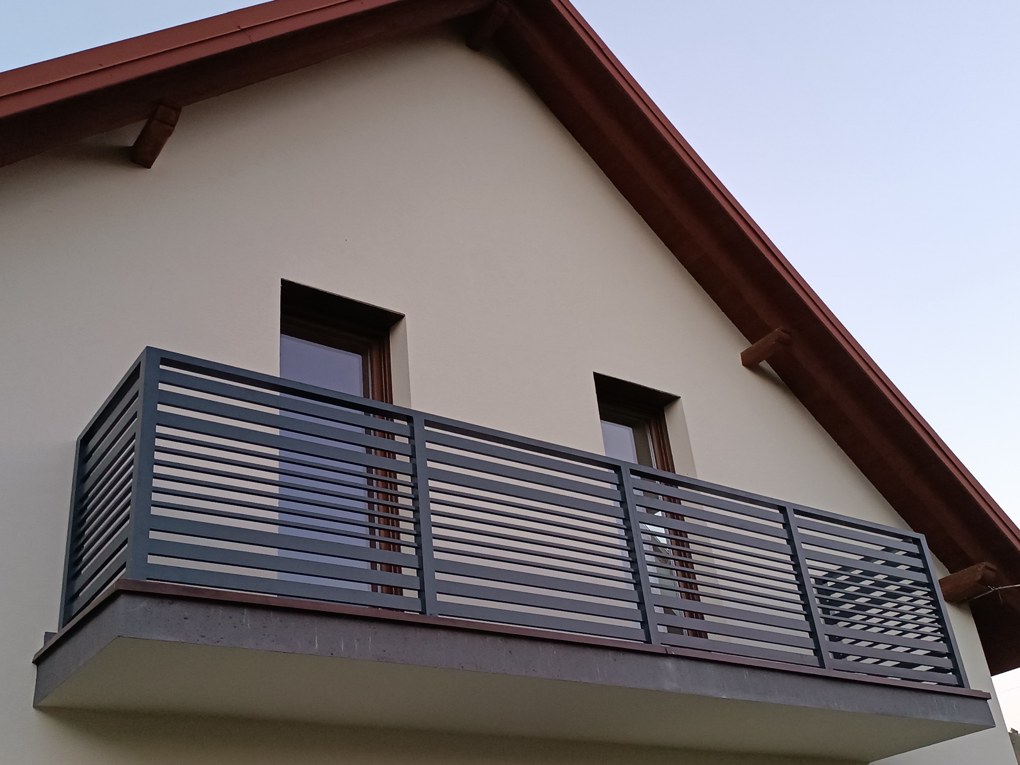 Nowoczesna, pozioma balustrada metalowa w kolorze grafitowym na balkonie domu jednorodzinnego. Widoczne dwa okna i fragment dachu z brązowym wykończeniem.