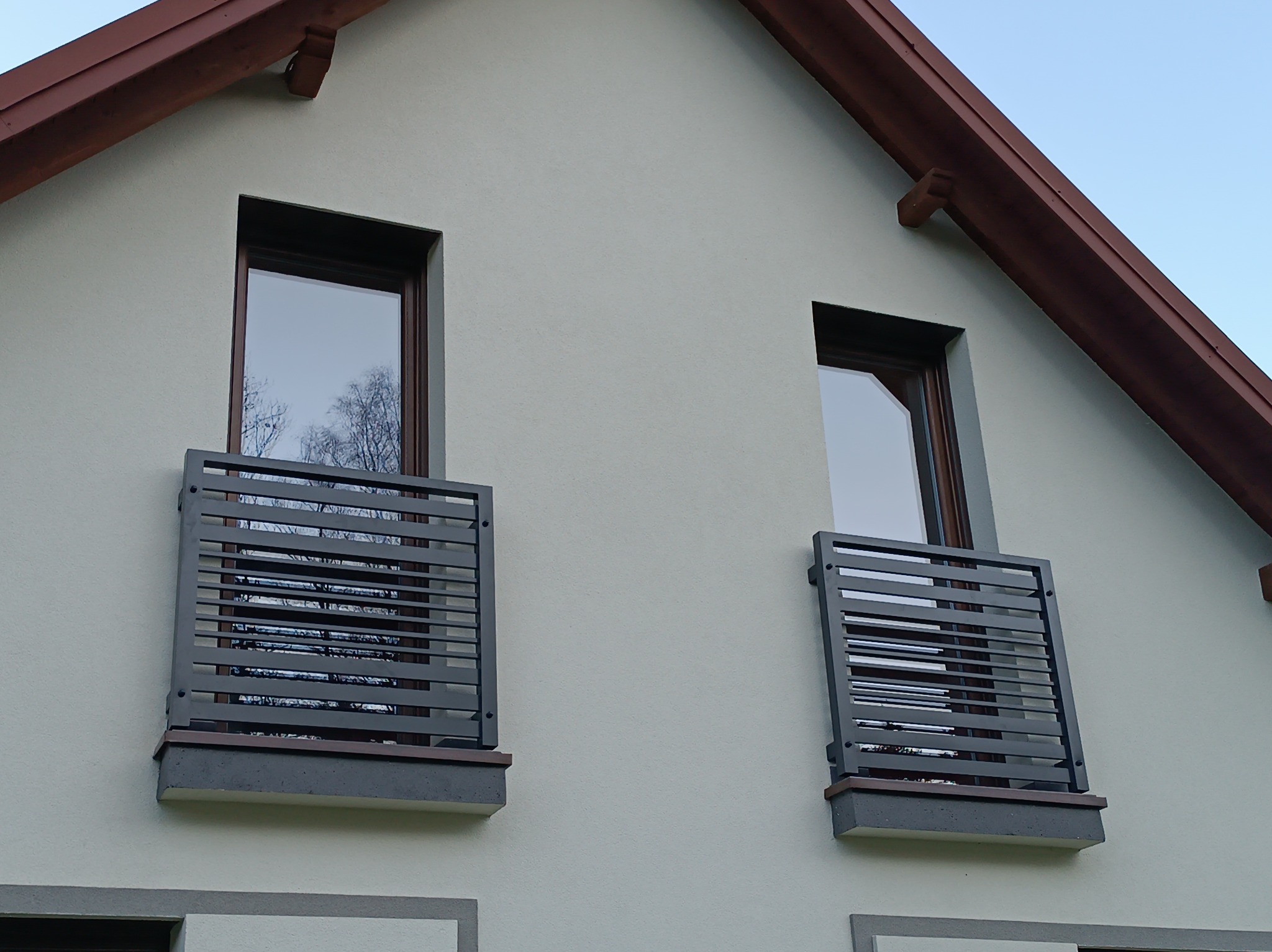 Elewacja domu z dwoma oknami balkonowymi zabezpieczonymi nowoczesnymi, poziomymi balustradami z metalowych profili w kolorze grafitowym. Widok z dołu, błękitne niebo.