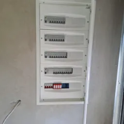 Rozdzielnica elektryczna ETI w ścianie z widocznymi zabezpieczeniami różnicowoprądowymi i nadprądowymi. Otwarta osłona skrzynki. Szara, surowa ściana w tle.