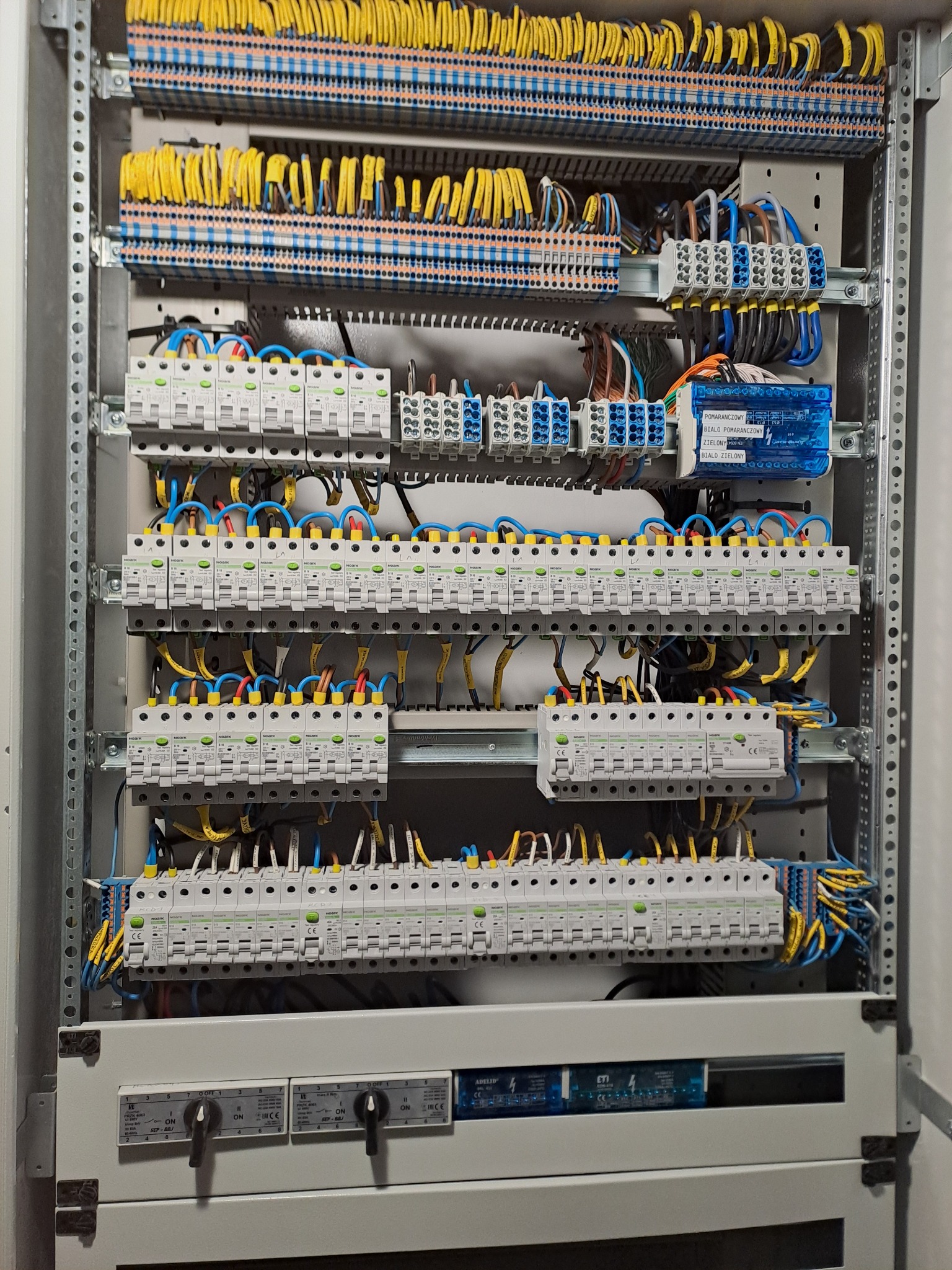 Rozdzielnia elektryczna z zabezpieczeniami, przewodami w kolorach żółtym i niebieskim, zamontowana w metalowej obudowie. Widoczne bezpieczniki i oznaczenia.