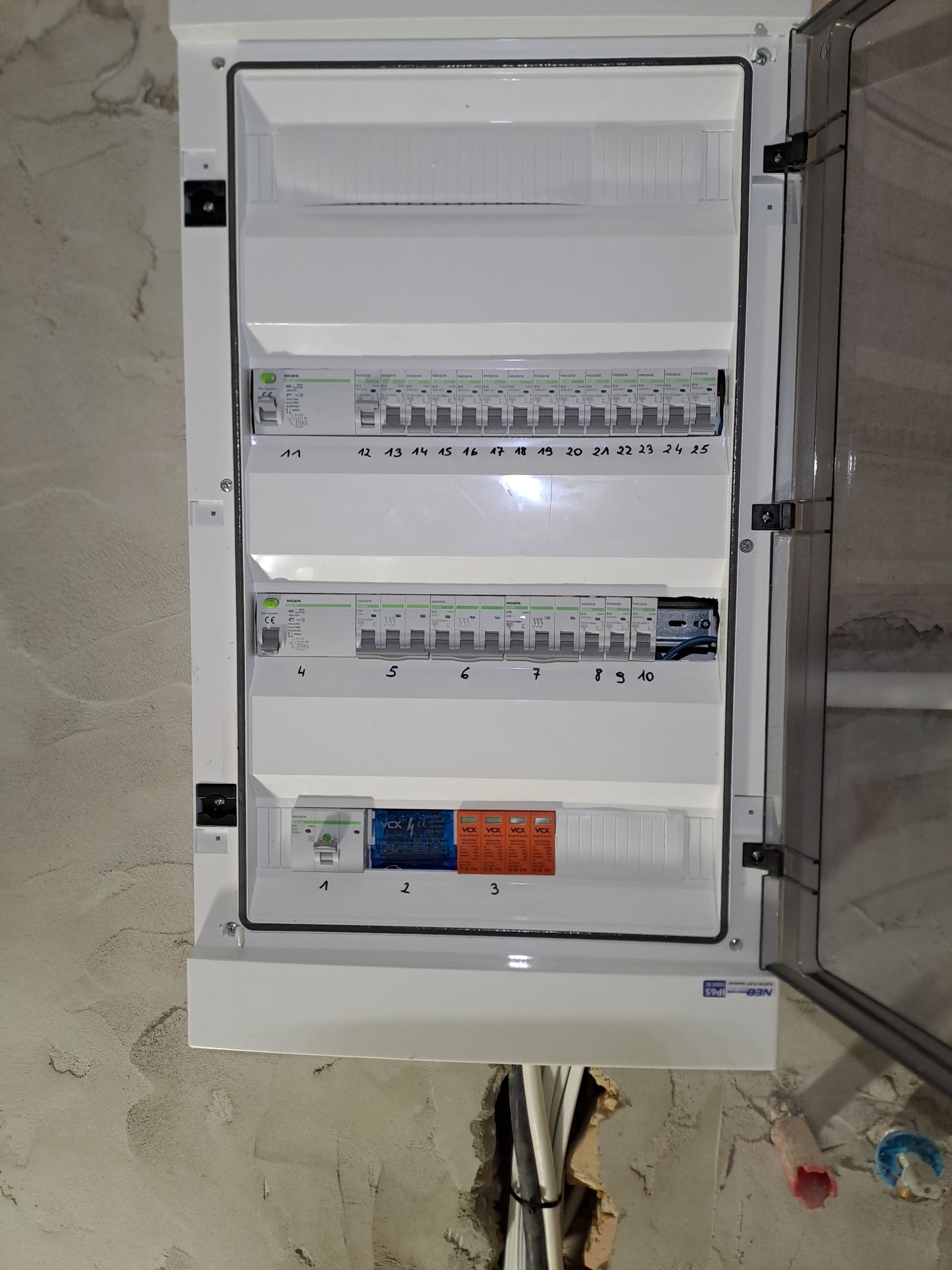 Rozdzielnica elektryczna natynkowa, z otwartymi drzwiczkami, widoczne bezpieczniki i numeracja. Ściana surowa, instalacja w peszlach.