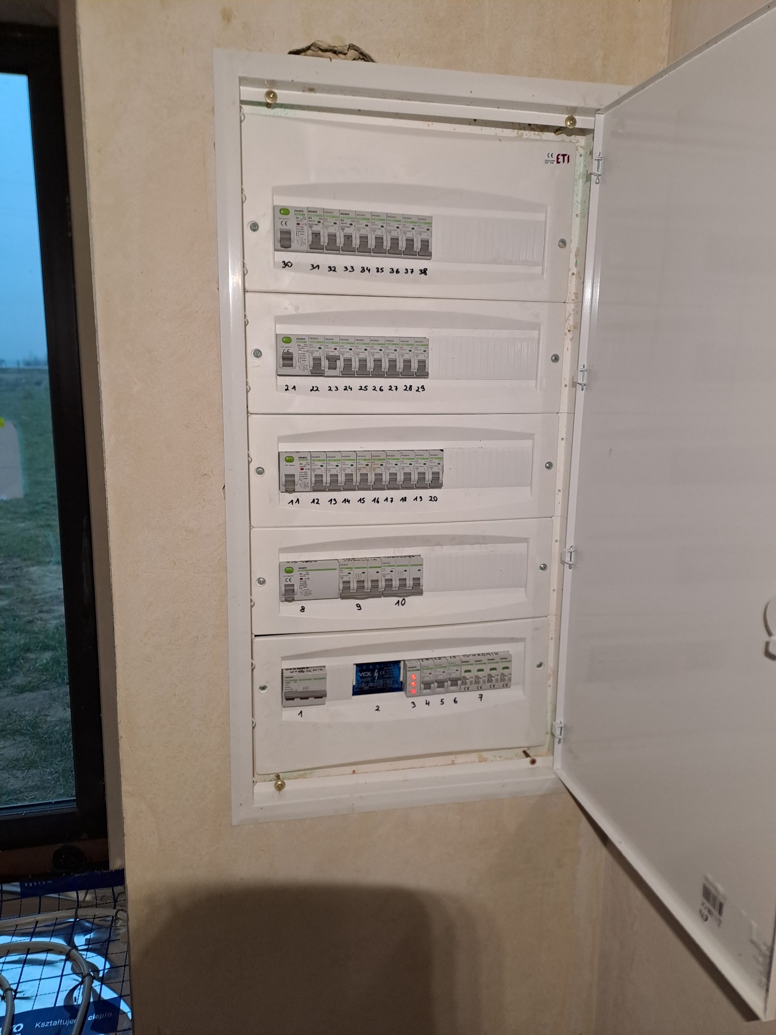 Rozdzielnica elektryczna z bezpiecznikami, widok z bliska. Otwarta obudowa ukazuje liczne zabezpieczenia i oznaczenia. Instalacja w nowym budynku, widok na okno.