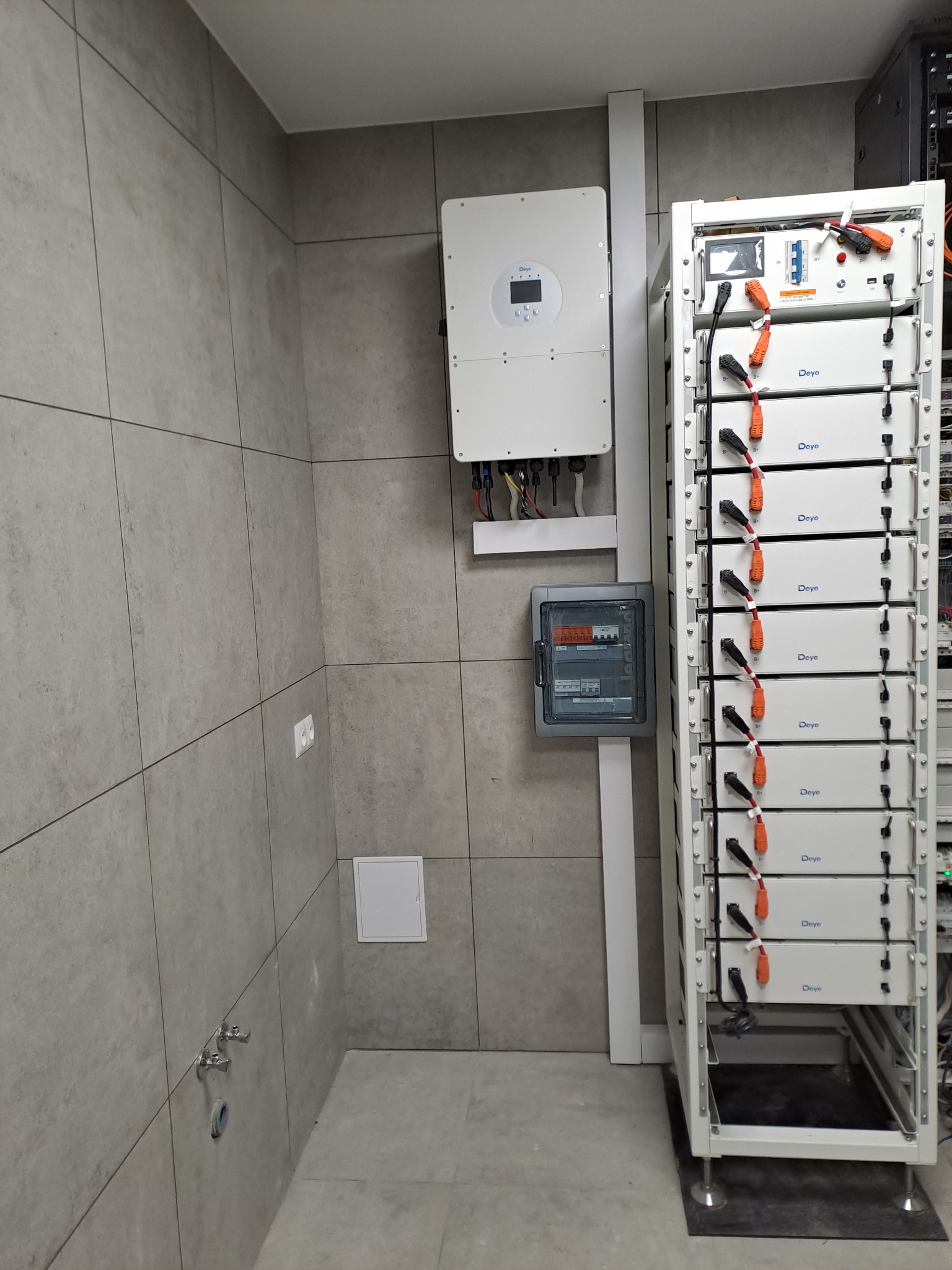 Biały inwerter solarny i bateria na ścianie z szarych płytek, obok metalowa szafka z akumulatorami. Widoczna skrzynka z bezpiecznikami i kable.