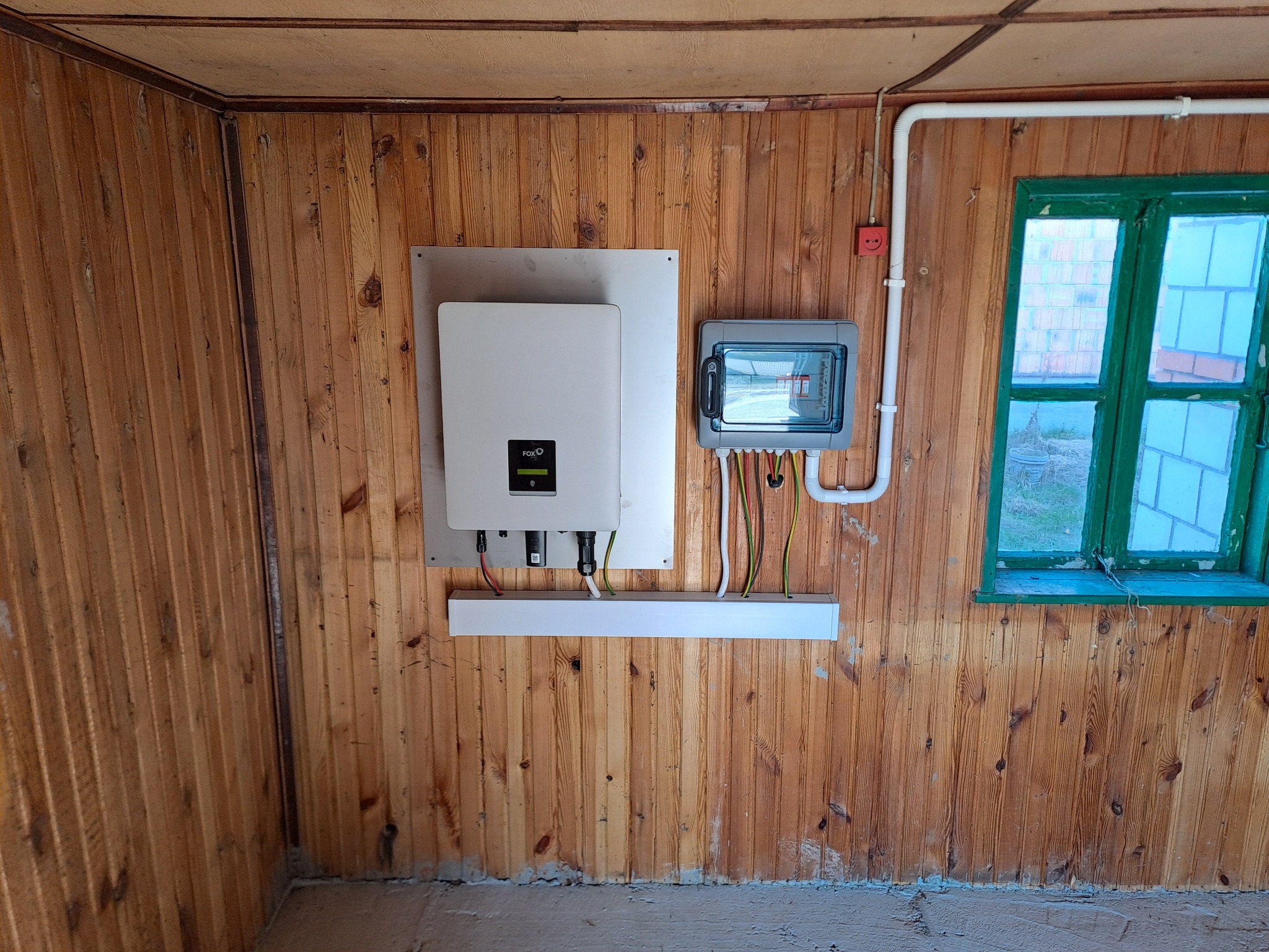 Zamontowany inwerter fotowoltaiczny FoxEss na drewnianej ścianie, obok skrzynka elektryczna i okno. Widoczne przewody i instalacja elektryczna.