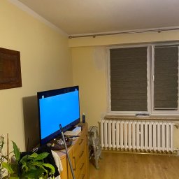 Grzegorz Sikora - Pokój z żółtymi ścianami, grzejnikiem i oknem z żaluzjami, fragment ściany przygotowany do malowania, drabina oparta o ścianę, meble zagracone przedmiotami, w rogu kwiat doniczkowy.