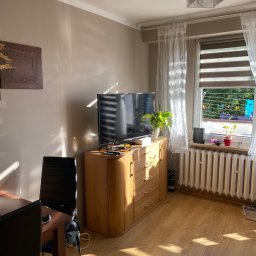 Grzegorz Sikora - Pokój dzienny z drewnianą komodą pod telewizorem, drewnianą podłogą i grzejnikiem pod oknem, na którym suszą się ubrania.